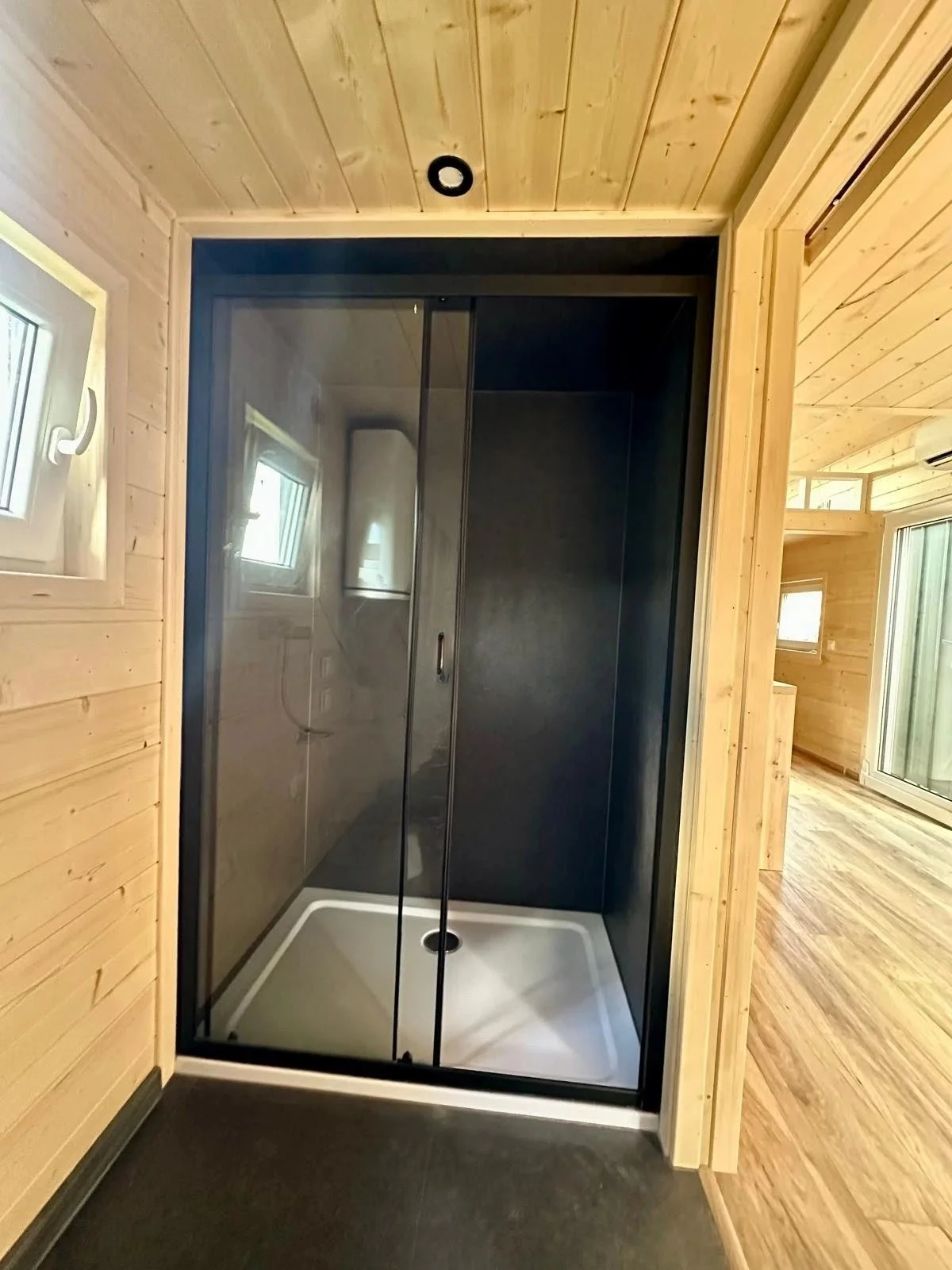 Ein moderner Duschbereich mit Glasschiebetüren, schwarzen Wänden, einem weißen Duschboden und einer kleinen Fenster in einem Holzhaus.
