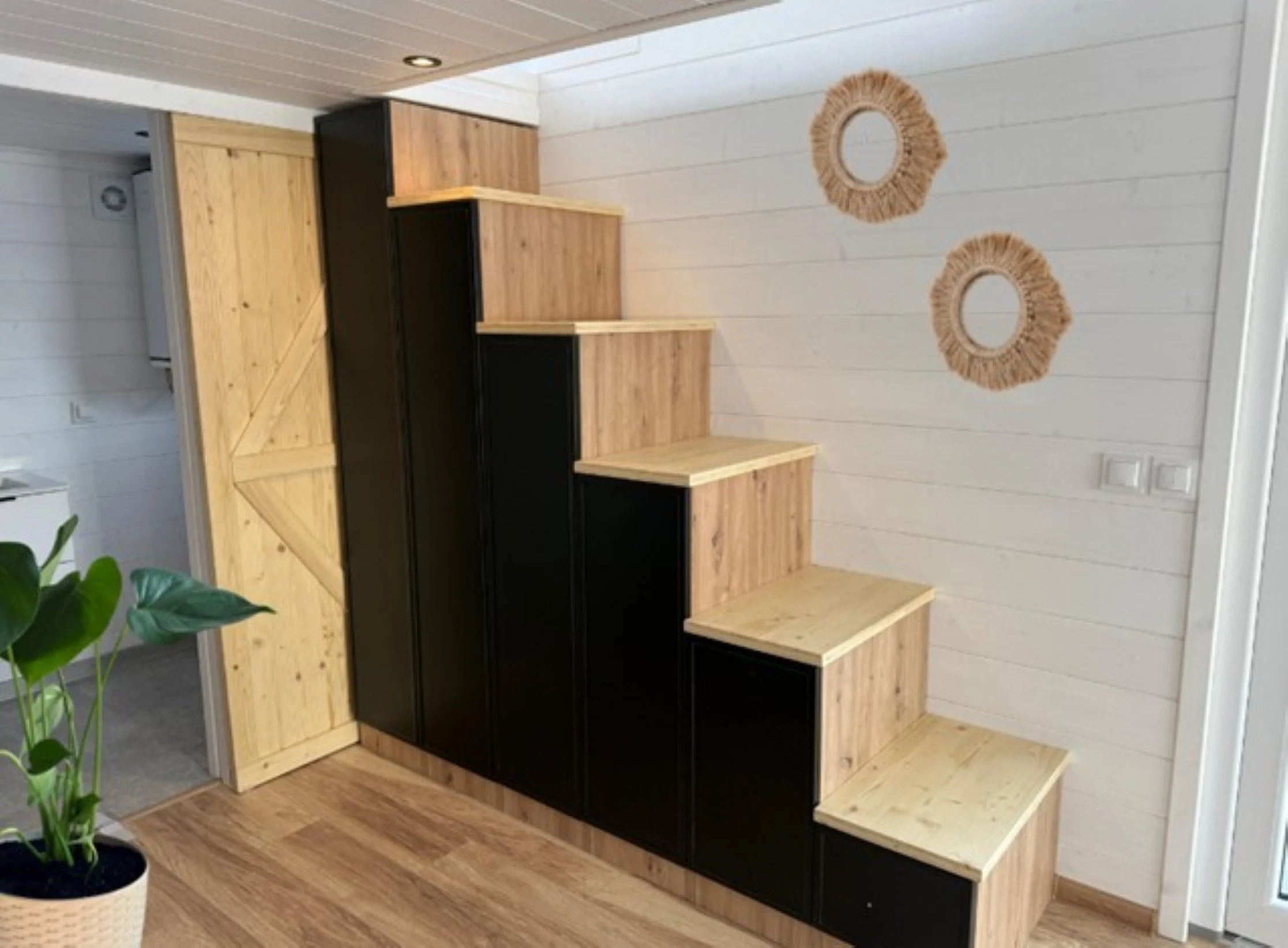 Treppenschrank mit schwarzer und natürlicher Holzfarbe, vor weißer Wand mit zwei runden Spiegeln, in einem Raum mit Holzfußboden und einer Pflanze in einem weißen Top.