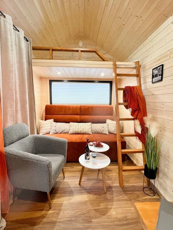 Gemütliches Wohnzimmer mit einem Betthaupt, einer kleinen Leseecke und einem Klettertreppe, die zu einem offenen Schlafbereich führt. Holzverkleidung an den Wänden und Decken, ein Fenster mit Rollos, ein Fernseher und Dekorationen.