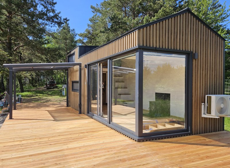 Moderne Holzhütte mit großen Glasfenstern und Holzdeck im Grünen. Auf der rechten Seite befindet sich eine Klimaanlage.