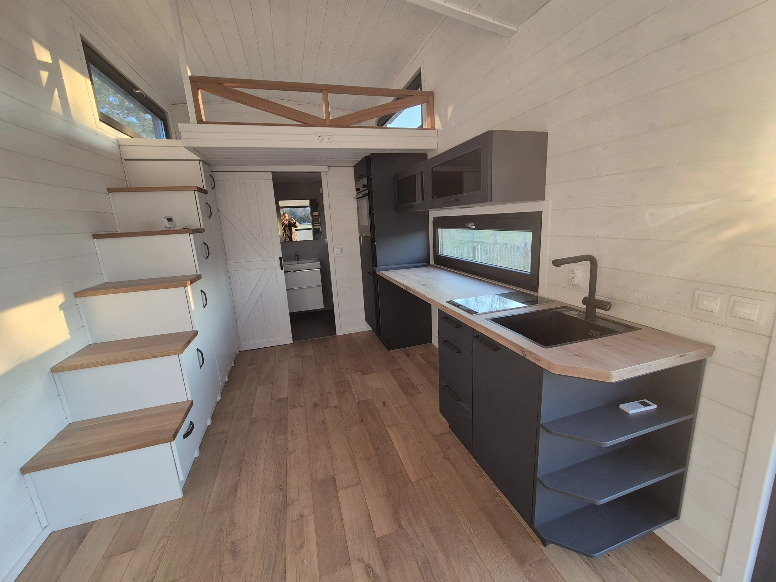 Moderne Küche mit weißen Wandvertäfelung, schwarzen Küchenschränken, Holzarbeitsplatten, großen Fenstern, zusammengeklappten weißen Treppen und einem Blick in eine kleine Waschküche.