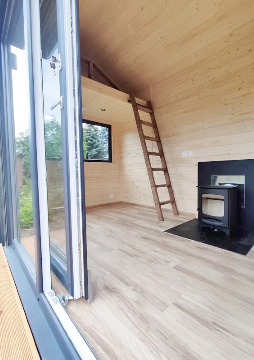Innenraum eines Holzhauses mit einem Kamin, einer Leiter zu einer offenen oberen Ebene, einem Fenster und einer glatten Holzdecke.