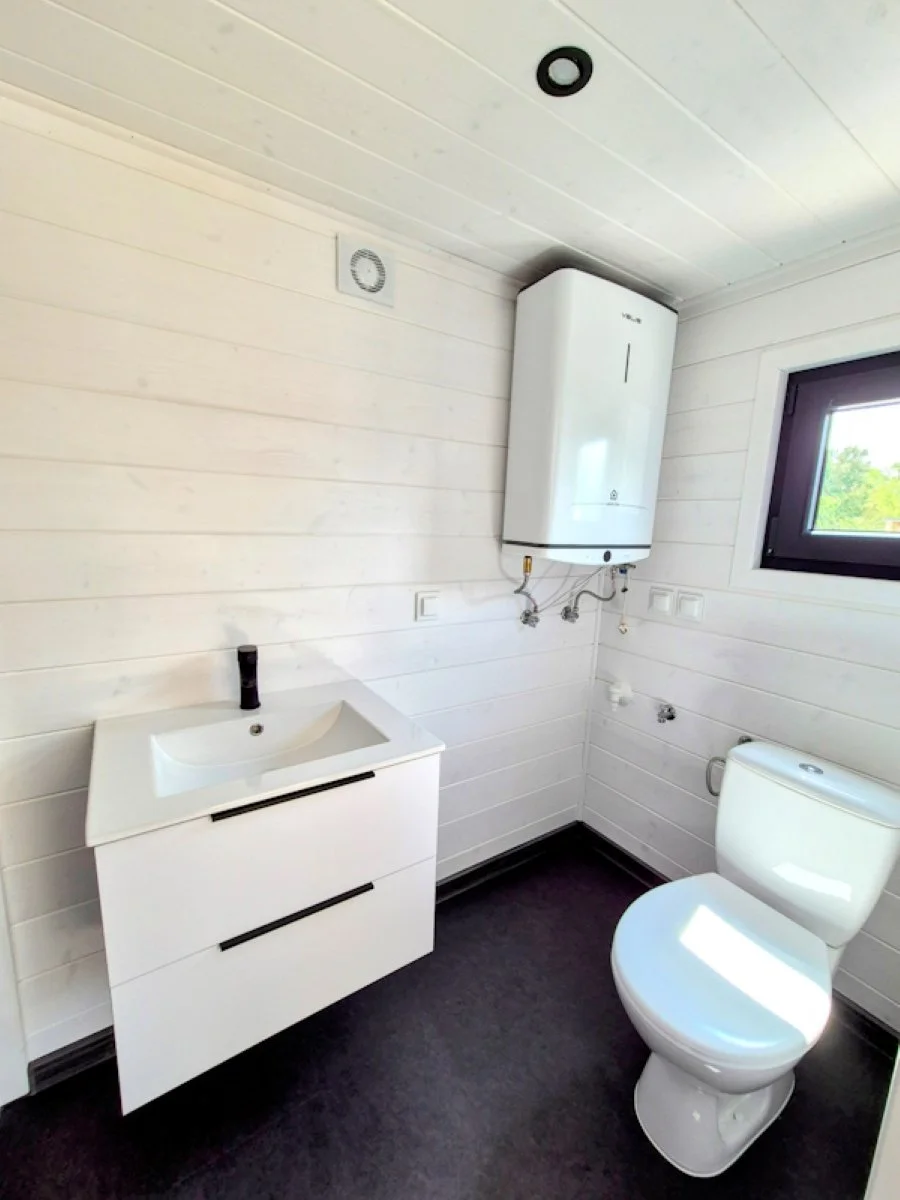 Ein modernes Badezimmer mit weißem Waschtisch, einer Toilette, einem Wasserboiler an der Wand und einem Fenster.