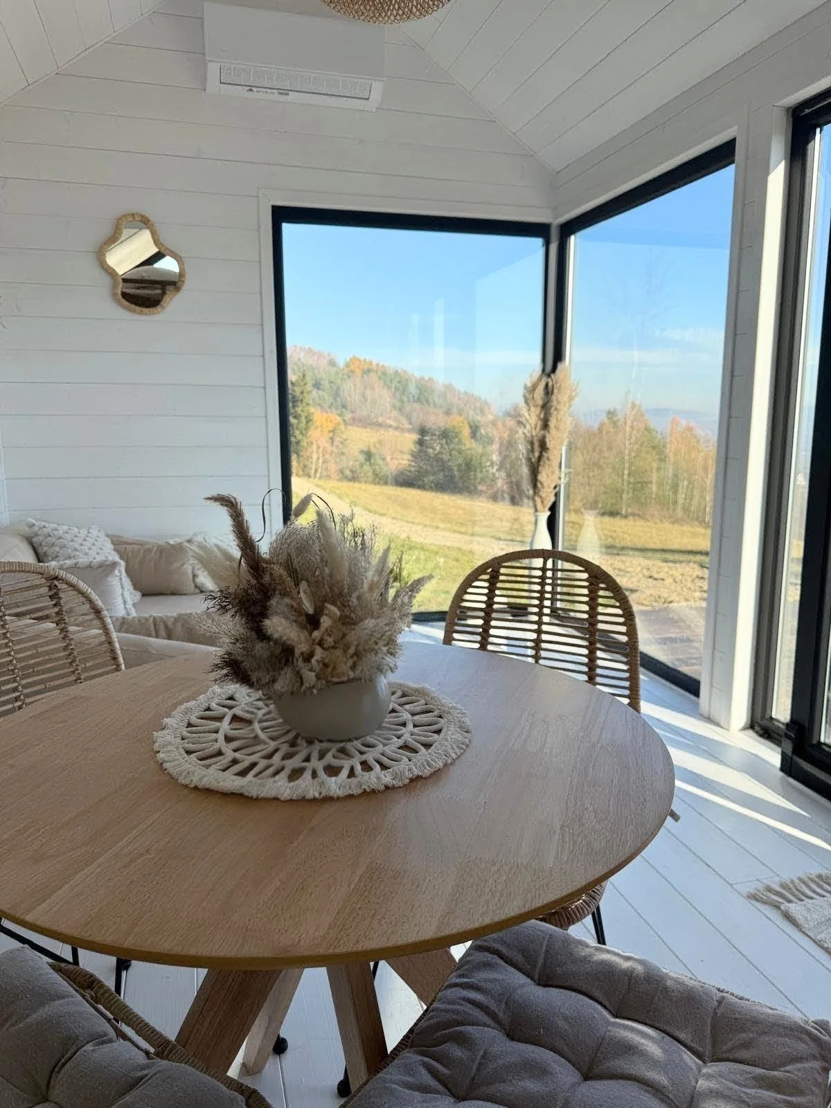 Moderner Wintergarten mit großen Fenstern, Blick ins Grüne, runder Holztisch mit Baumwoll-Tischdecke und Dekoration aus Federn, Rattan-Stühle, gemütliche Couch mit weißen Kissen, weiße Wände mit Spiegel, natürlicher Lichtschein.