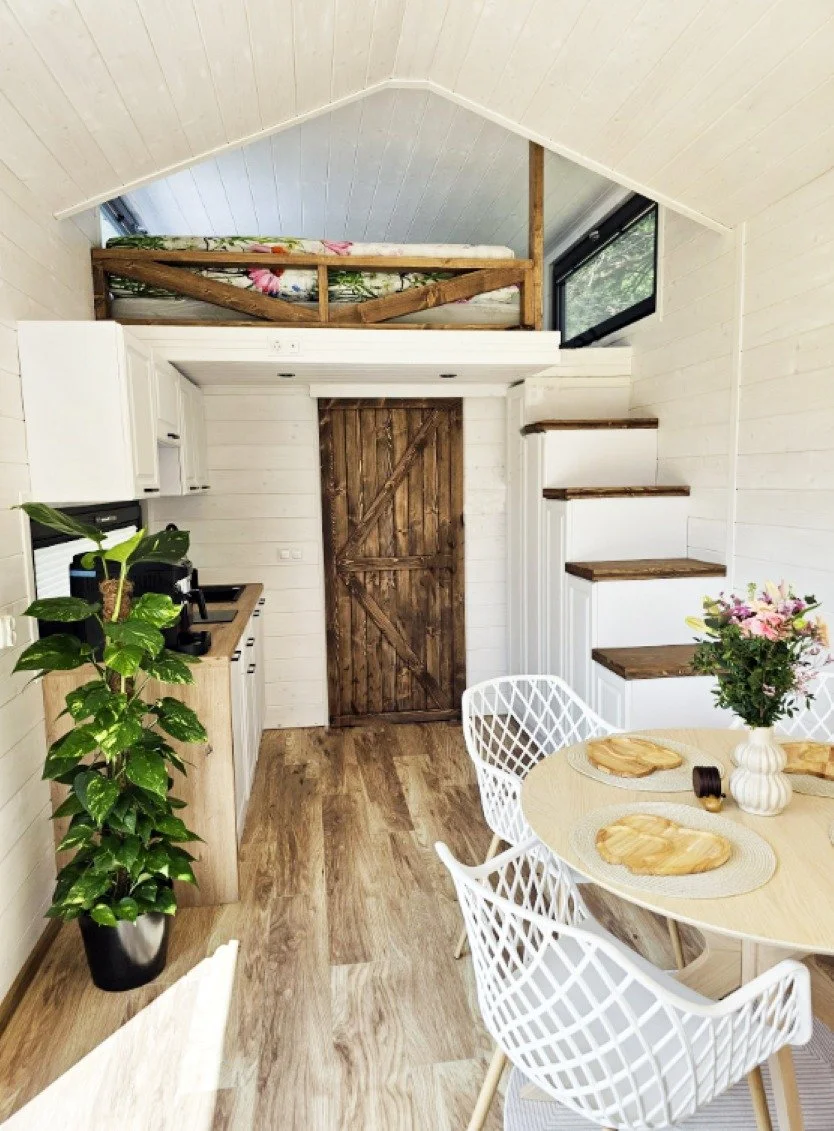 Interior eines kleinen, gut eingerichteten Hauses mit Küchenbereich, Essbereich und einem Loft-Schlafbereich. Weiße Wände, Holzboden und Holzdetails.