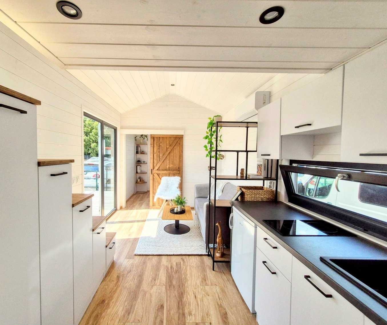 Moderner, heller Küchenraum mit weißen Schränken, schwarzen Griffen, schwarzen Kochplatten, Holzfußboden, Sicht auf einen Wohnbereich mit Sofa, Sessel und Tür aus Holz, viel Tageslicht durch große Fenster und Türen.