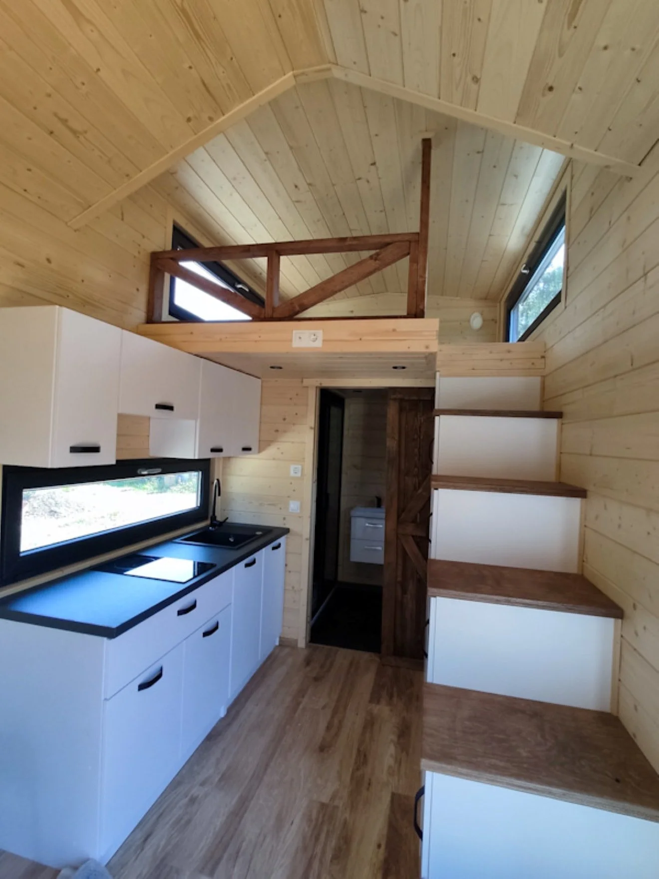 Ein kleiner Küchenraum mit weißen Schränken, einem schwarzen Herd, einem schwarzen Spülenbecken und Holzregalen. Die Wände und Decken sind aus Holz, mit kleinen hohen Fenstern, die natürliches Licht lassen, und einer Holztreppe auf der rechten Seite.