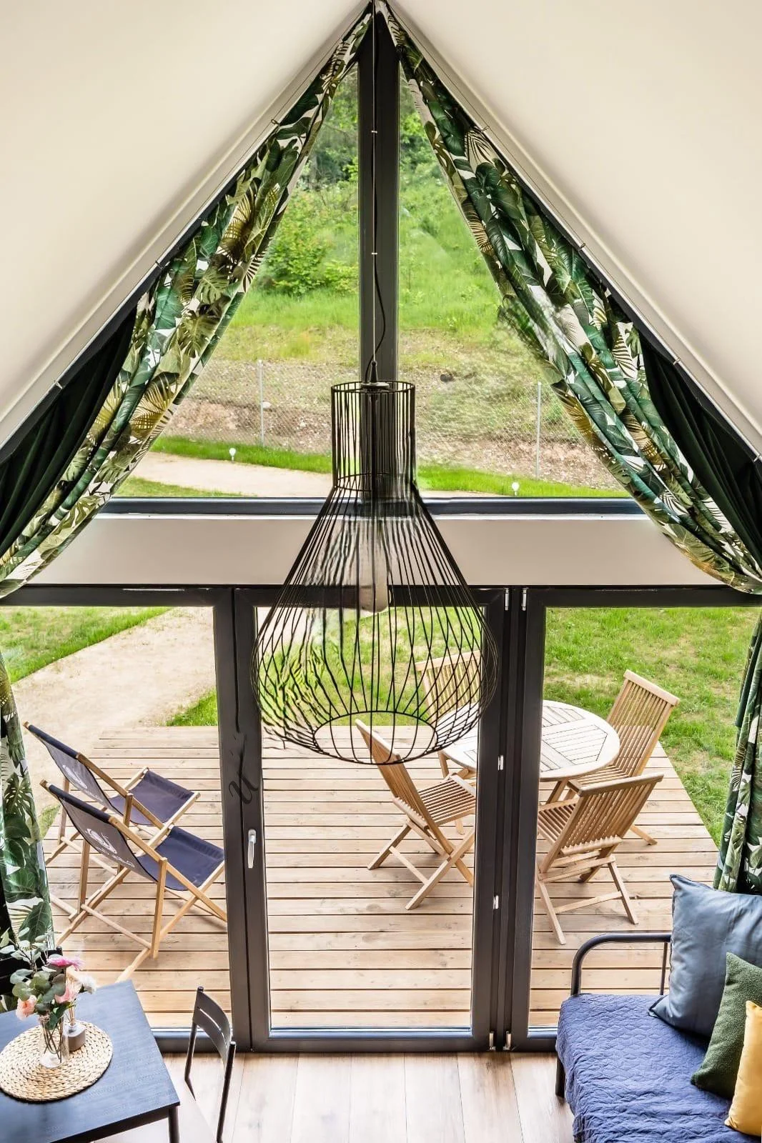 Innenaufnahme mit Blick auf eine offene Terrassentür, die zu einer Holzterrasse führt. Im Raum hängt eine schwarze Lampe und es sind Sitzgelegenheiten zu sehen, darunter ein Sofa mit Kissen. Die offene Tür ist mit grünen, blättrigen Gardinen dekorier