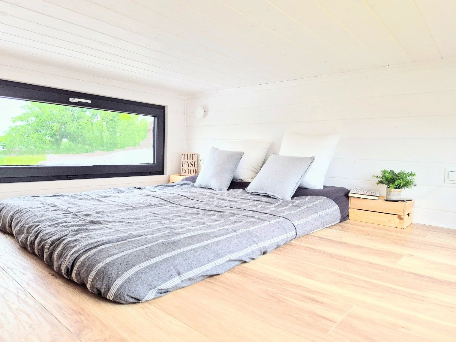 Schlafzimmer mit niedrigem Bett auf dem Boden, grauem Bettzeug, weißen und grauen Kissen, seitlichen Nachttischen mit Büchern und Pflanzen, großes Fenster mit Blick auf grüne Bäume, weiße Wand und Holzdecke.