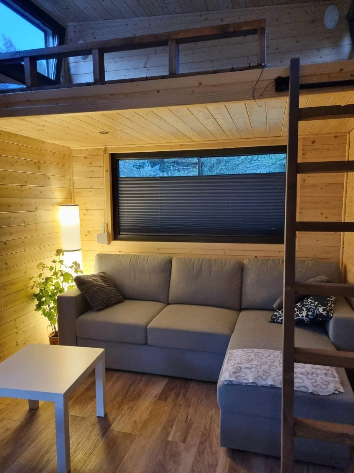Gemütliches Wohnzimmer mit Holzvertäfelung, grauem Sofa, kleinem weißen Tisch, einer Stehlampe, einem Fenster mit Rollladen und einer oberen Etage mit Treppe aus Holz.