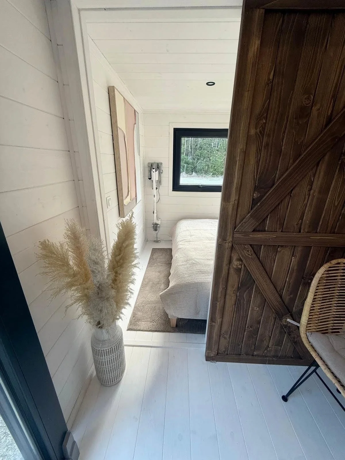 Innenaufnahme eines Kamins, mit einem weißen Sofa, einem Fenster, einem Vasen mit Trockenblumen, einem Holzwandtor und einem Stuhl.