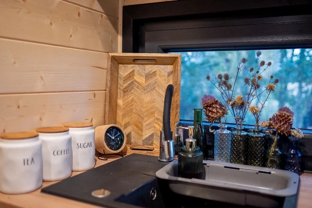 Küchenrolle und dunkler Wasserhahn über einer Spüle, auf der Fensterbank befinden sich mehrere Vasen mit getrockneten Blumen, im Hintergrund ein Fenster mit Blick nach draußen, Küchenutensilien wie eine Uhr, drei Keramikdosen mit Holzdeckeln für Tee,