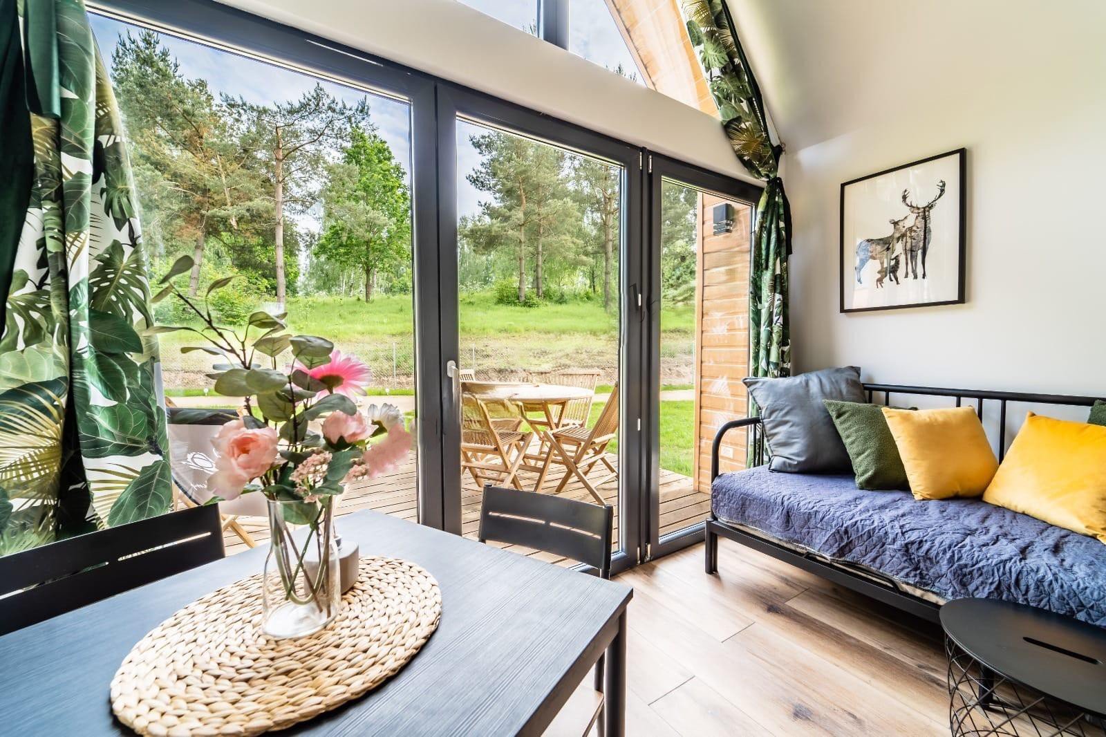 Innenraum eines Wohnzimmers mit großen Glastüren, Gartenmöbeln im Garten und einer Couch mit bunten Kissen.