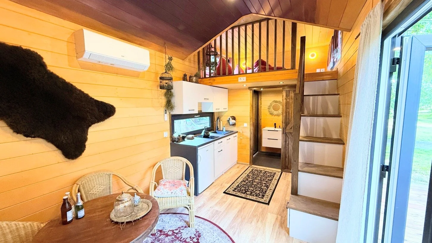 Innenaufnahme einer kleinen Holzhausküche mit Loft, es gibt ein kleines Waschbecken, einen Tisch mit Stühlen, eine Schrankwand, Stufen zum Loft, Holzverkleidung, und ein Fenster mit Blick nach draußen.