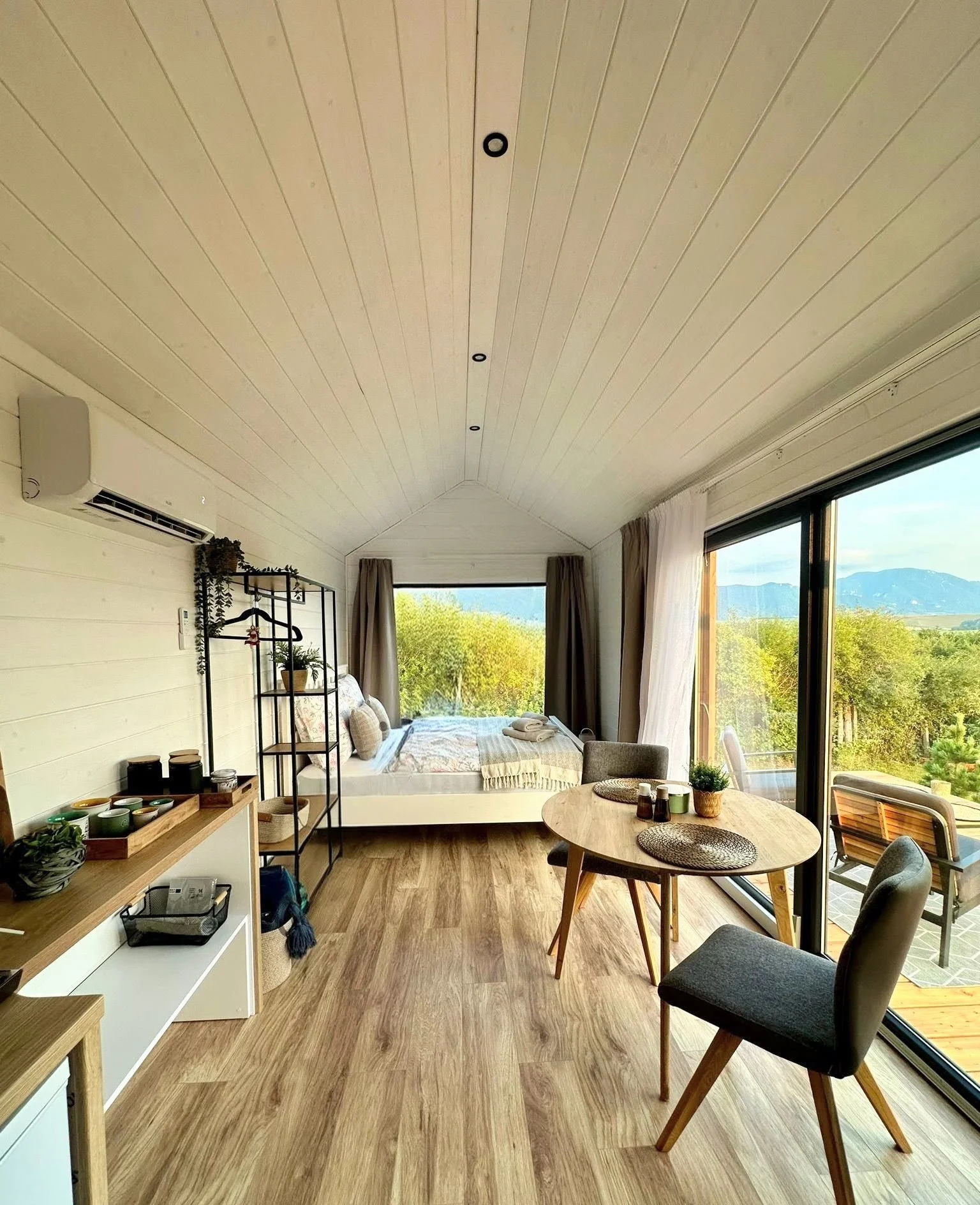 Ein gemütliches Zimmer mit Holzboden, Doppelbett, Esstisch und Stühlen, großer Fensterfront mit Blick auf Bäume und Berge, fürnatmatischer Stil, viel Tageslicht und moderner Einrichtung.