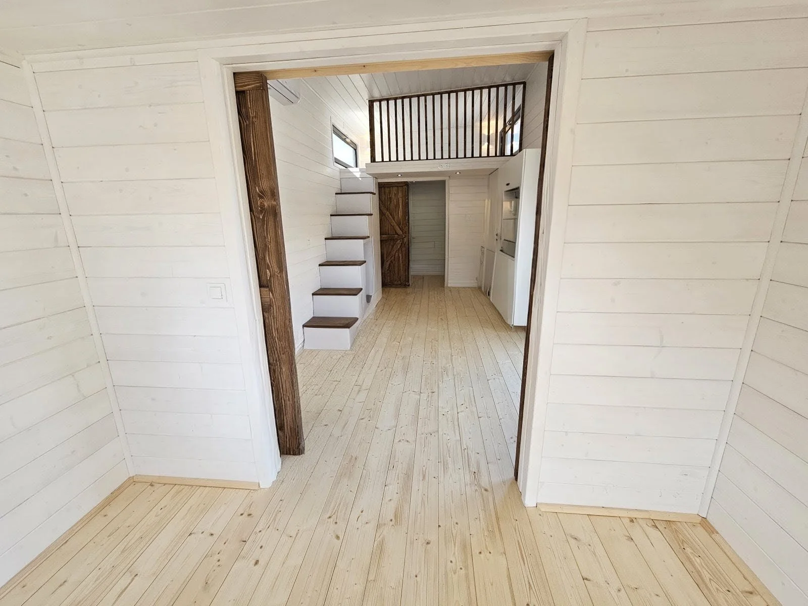 Innenaufnahme eines hellen, modernen Hauses mit Holzboden, weißen Wänden und einer Treppe mit dunklen Stufen, die nach oben führt.