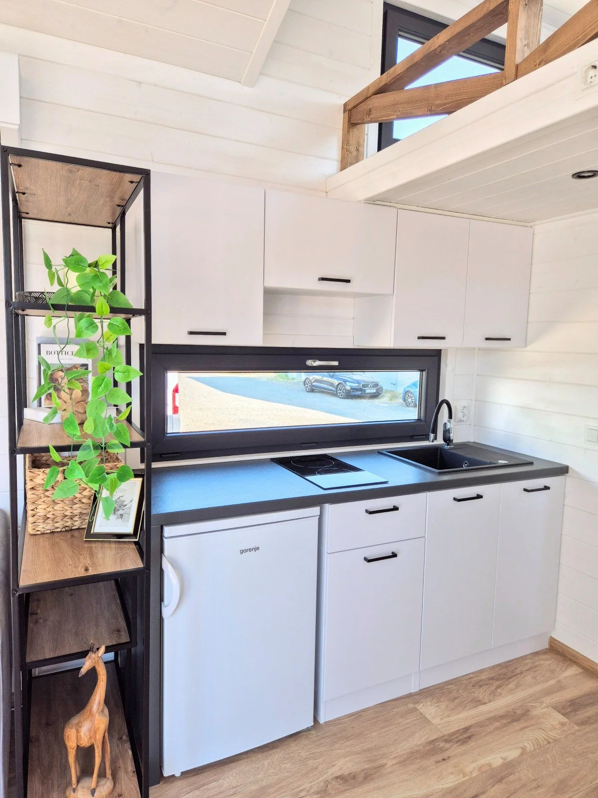 Küchenzeile mit weißen Schränken, schwarzer Arbeitsplatte, Wasserhahn, Spüle, kleiner Kühlschrank und ein Fenster oberhalb der Arbeitsfläche. Es gibt ein offenes Regal mit Pflanzen und Dekorationsgegenständen auf der linken Seite.