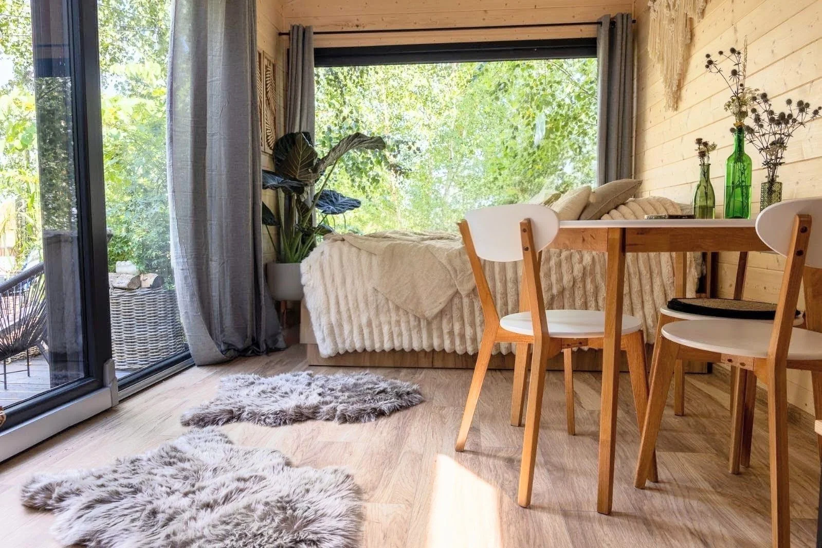 Gemütliches Wohn- und Esszimmer mit Holzdekoration, einem großen Fenster, Pflanzen, einem Sofa mit Decke, einem Tisch mit drei weißen Stühlen, und einem Balkon.