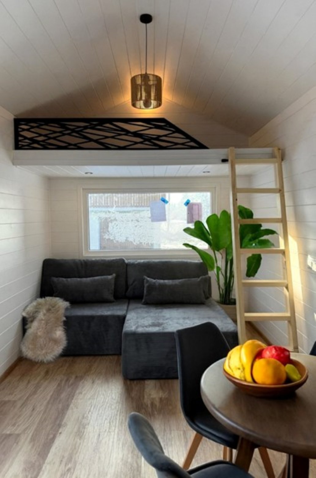 Ein modernes Wohnzimmer mit einer hohen Decke, Sofakissen, einem Liegesofa, einer Pflanzen, einem Fenster, einem Esstisch mit einer Obstschale, einer Leiter und einem erhöhten Schlafbereich.