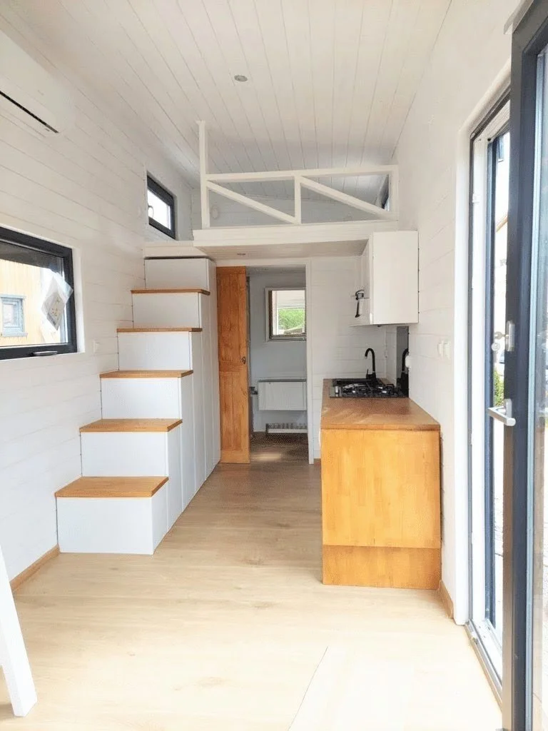Kleine Küche mit weißen Wänden, Holzboden und einem Mini-Kochbereich auf der rechten Seite. Es gibt eine Treppe mit weißen Stufen, die nach oben führt, und ein Fenster auf der linken Seite.