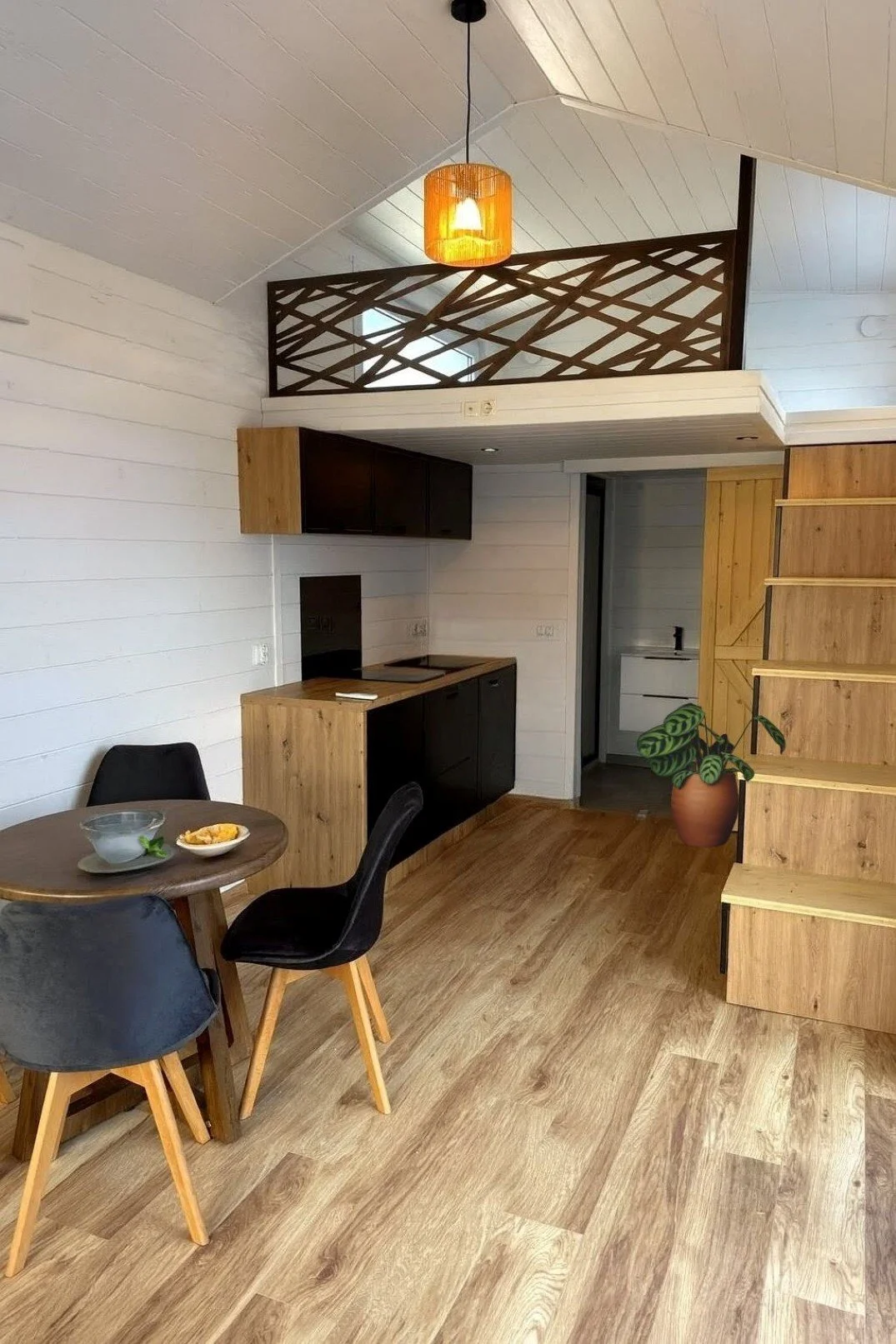Innenaufnahme eines Hauses mit Holzboden, weiß gestrichenen Wänden, einer kleinen Küchenzeile mit schwarzen Schränken, einem runden Esstisch mit drei schwarzen Stühlen, einer Treppe aus Holz und einem Balkon mit Holzgeländer, sowie einer großen Topfp