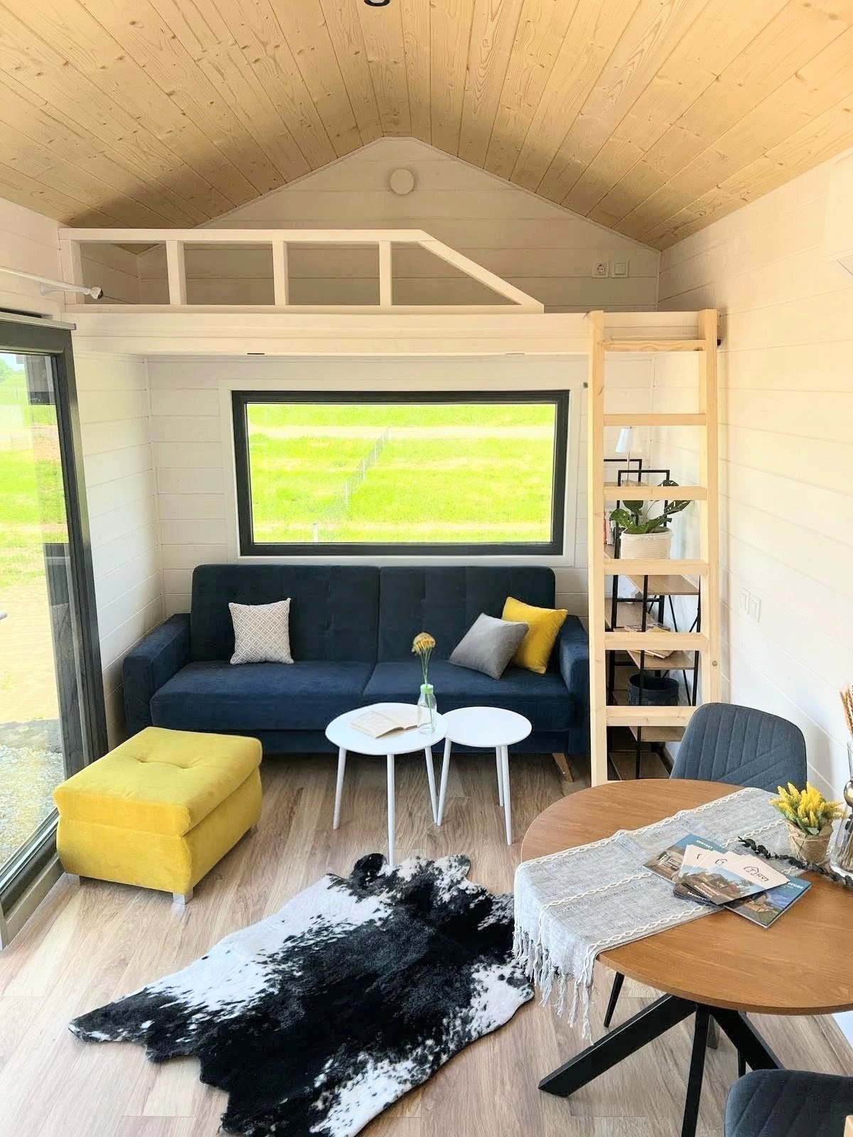 Ein modernes Wohnzimmer mit einer dunkelblauen Couch, bunten Kissen, einem gelben Hocker, zwei weißen Beistelltischen, einem Teppich, einem runden Holztisch, einem schwarzen Stuhl und einer Leiter, die zu einem Schlafboden führt, vor einem großen Fen