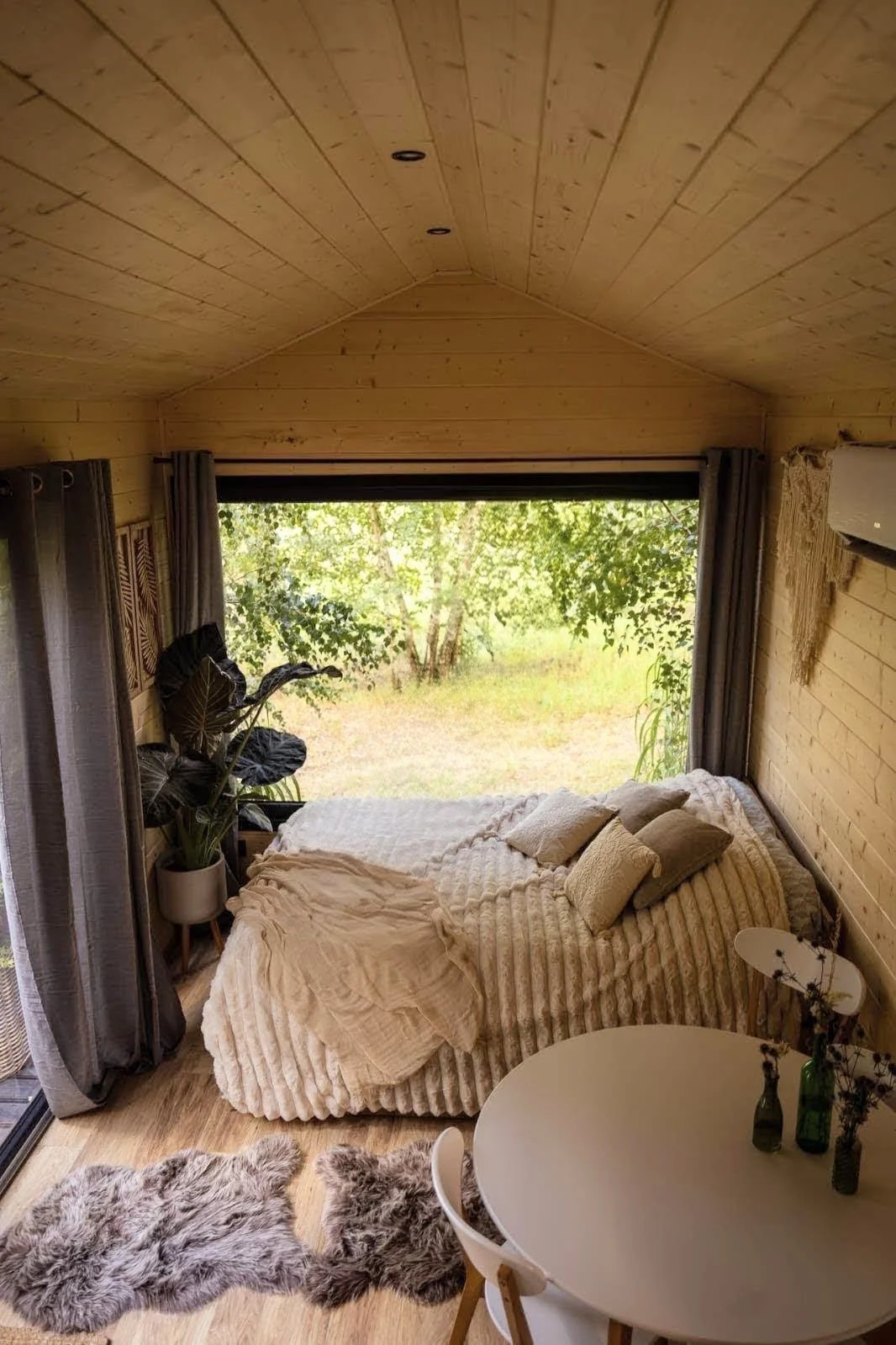 Ein gemütliches Schlafzimmer mit Holzwänden und -decken, einem großen Bett mit weißen Kissen und Decke, großem Fenster mit Blick ins Grüne, einer Pflanze in einer weißen Topf, einem runden Tisch mit Blumenvasen, und zwei flauschigen Teppichen auf Hol