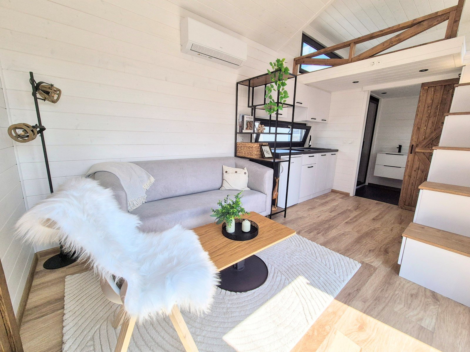 Gemütliches Wohnzimmer mit weißen Wänden, grauem Sofa, Holztisch mit Pflanzen, weißem Fellstuhl, offener Küche mit weißen Schränken und schwarzen Ablagen, Treppe mit weißen Stufen und Holzdetails, große Fenster mit natürlichem Licht.