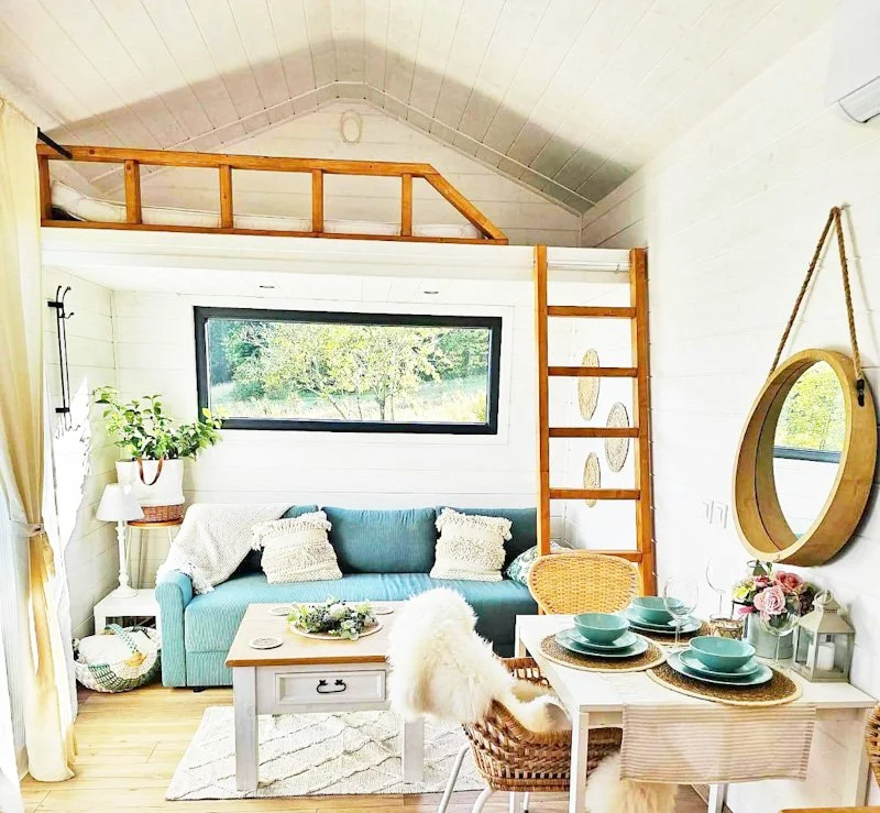 Helles Wohnzimmer mit blauem Sofa, weißen Kissen, Holztreppe, großen Fenstern, runder Spiegel, gedecktem Tisch mit Set von Tellern und Blumen, Pflanzen, und gemütlicher Dekoration.