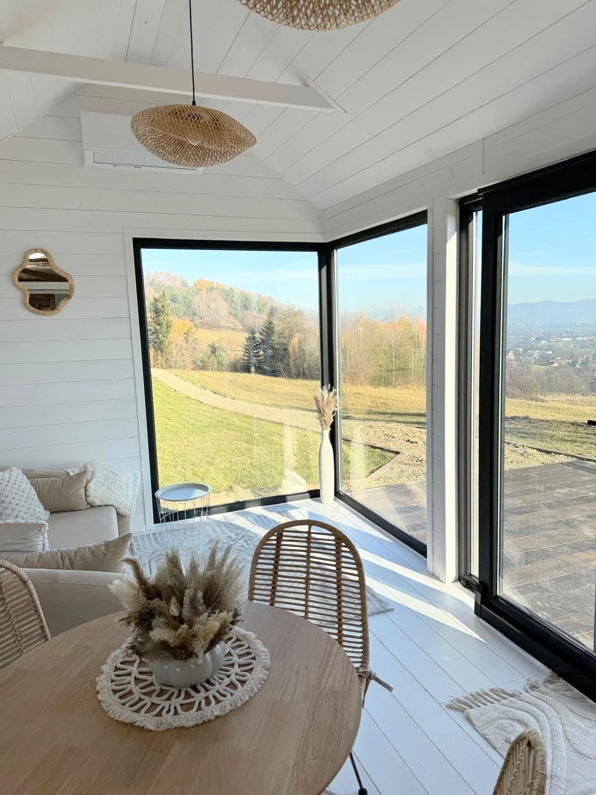 Ein heller, moderner Raum mit großen Fenstern, die einen Ausblick auf eine grüne, hügelige Landschaft bieten. Es gibt einen Rattanstuhl, ein Holztablett mit Dekorationen, weiße Wände und eine bequeme Couch. Die Einrichtung ist im skandinavischen Desi