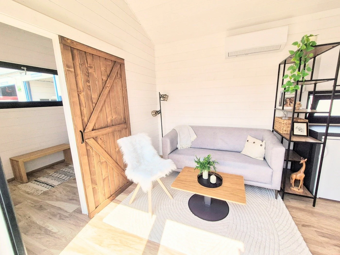 Gemütliches Wohnzimmer mit grauem Sofa, kleinen Kissen, dunklem Beistelltisch, Holz- und Stahlregal mit Dekorationen, Pappelscheife-Tür, Holzboden, Fenster und Wandklimaanlage.