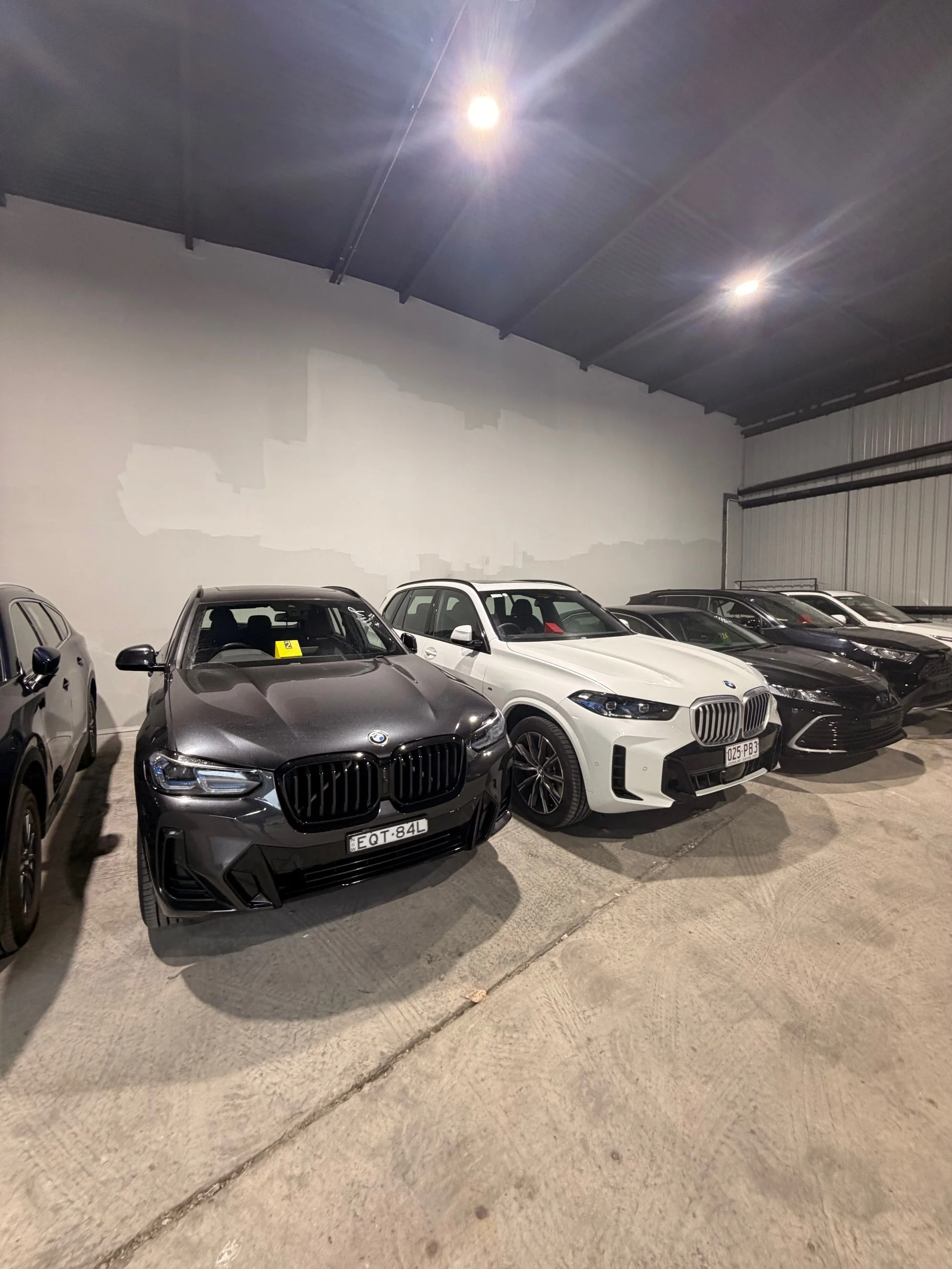 A row of luxury cars parked inside a well-lit garage with a concrete floor and a partially painted white wall.