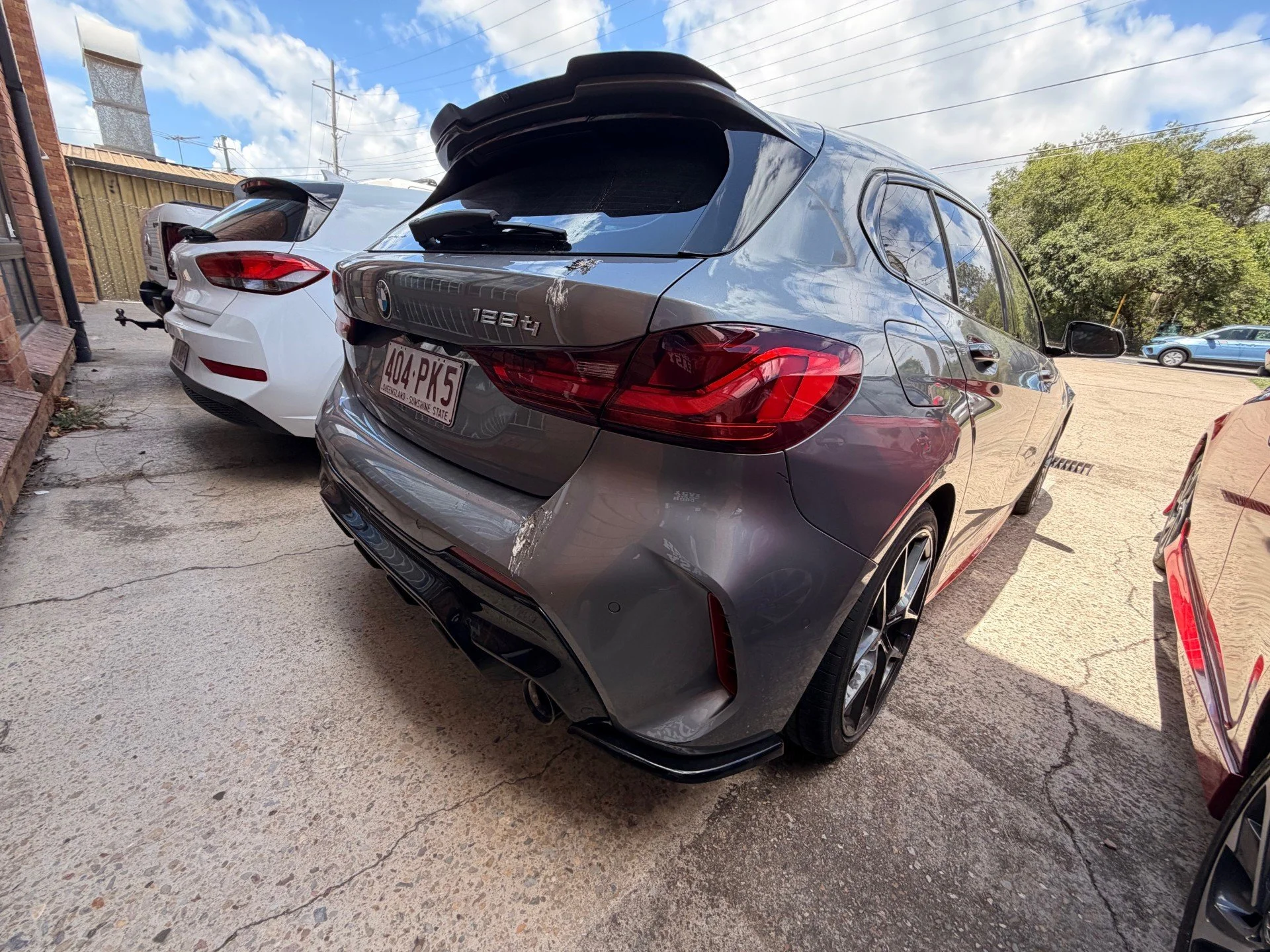 A black BMW 1284 parked in a lot, with a white vehicle and another car visible nearby, and a partly cloudy sky overhead.