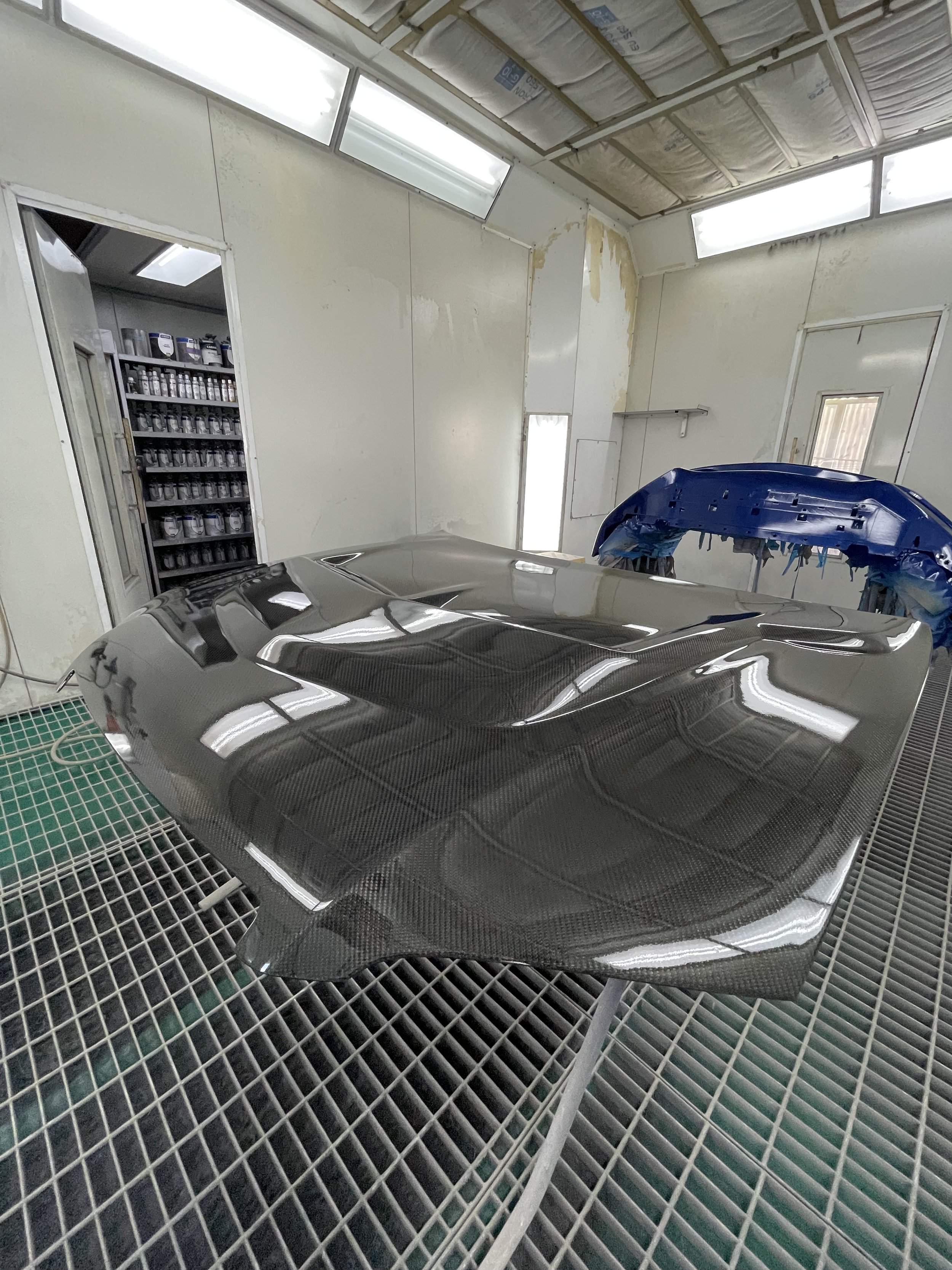 A carbon fiber car hood drying on a rack in a paint booth with white walls and a ceiling with fluorescent lights.