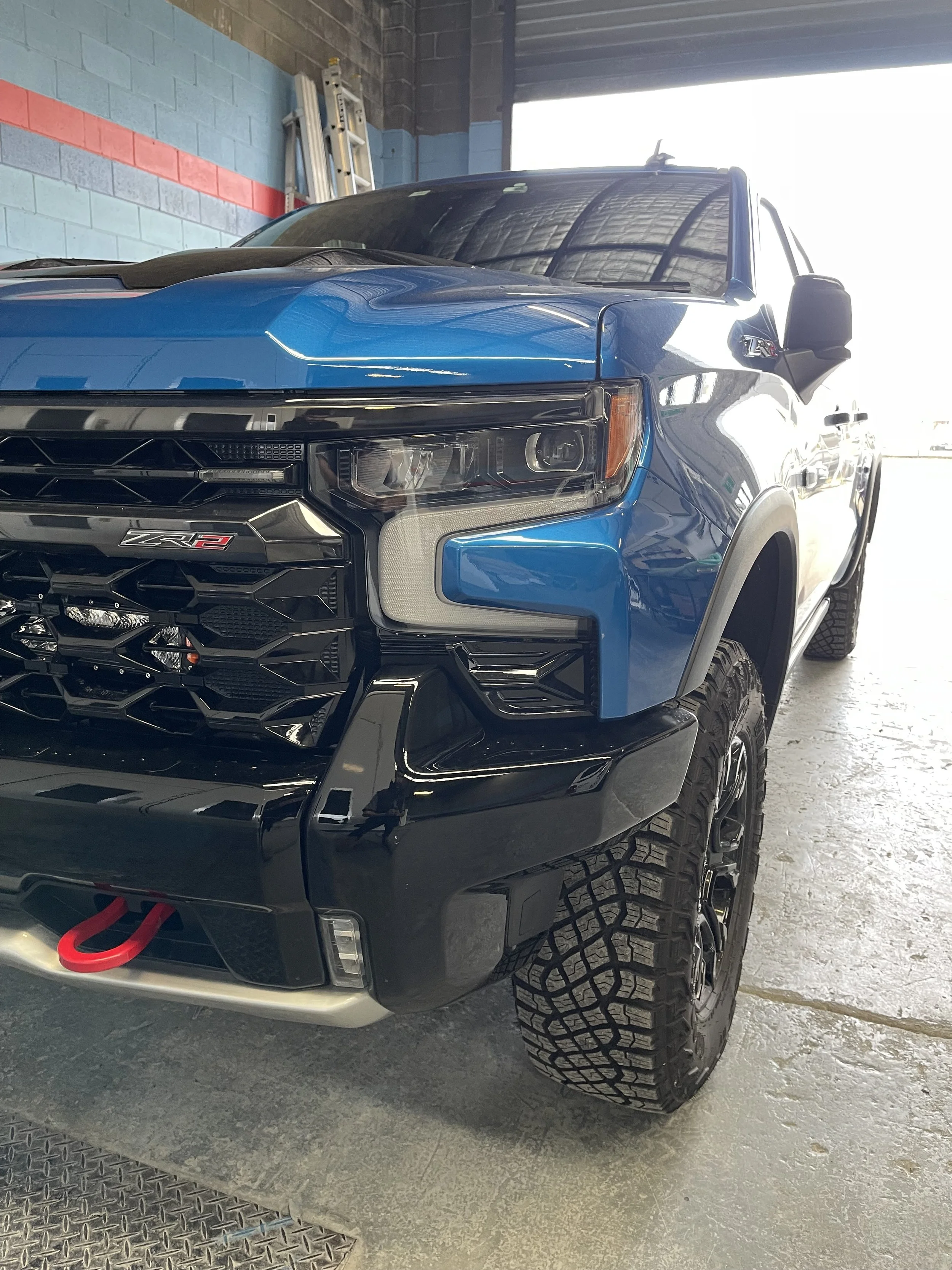 Front view of a blue and black pickup truck with off-road tires, parked inside a garage with a blue and gray painted wall.