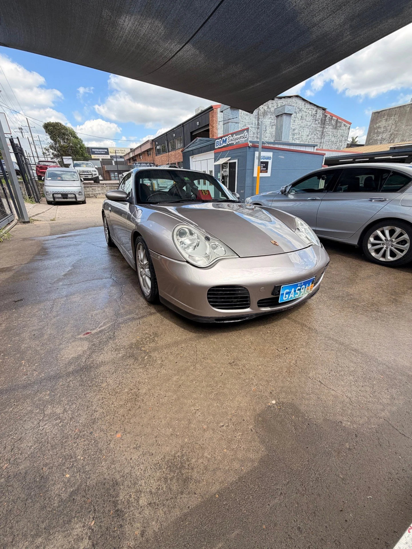 Silver Porsche sports car parked under a canopy at a car lot, with other cars and buildings in the background, and a partly cloudy sky overhead.