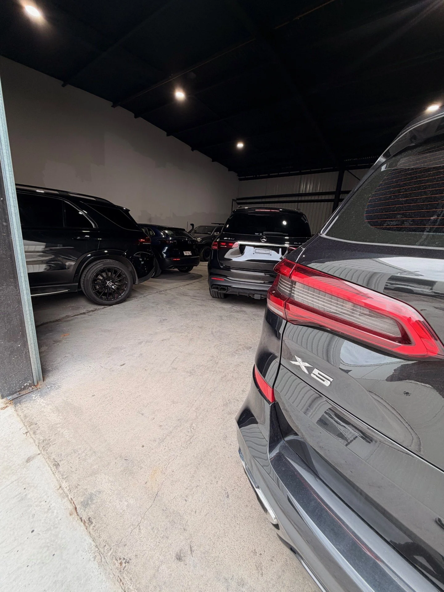 Inside a parking garage with multiple black and dark-colored cars, including an SUV and a sedan, with a concrete floor and artificial ceiling lights.