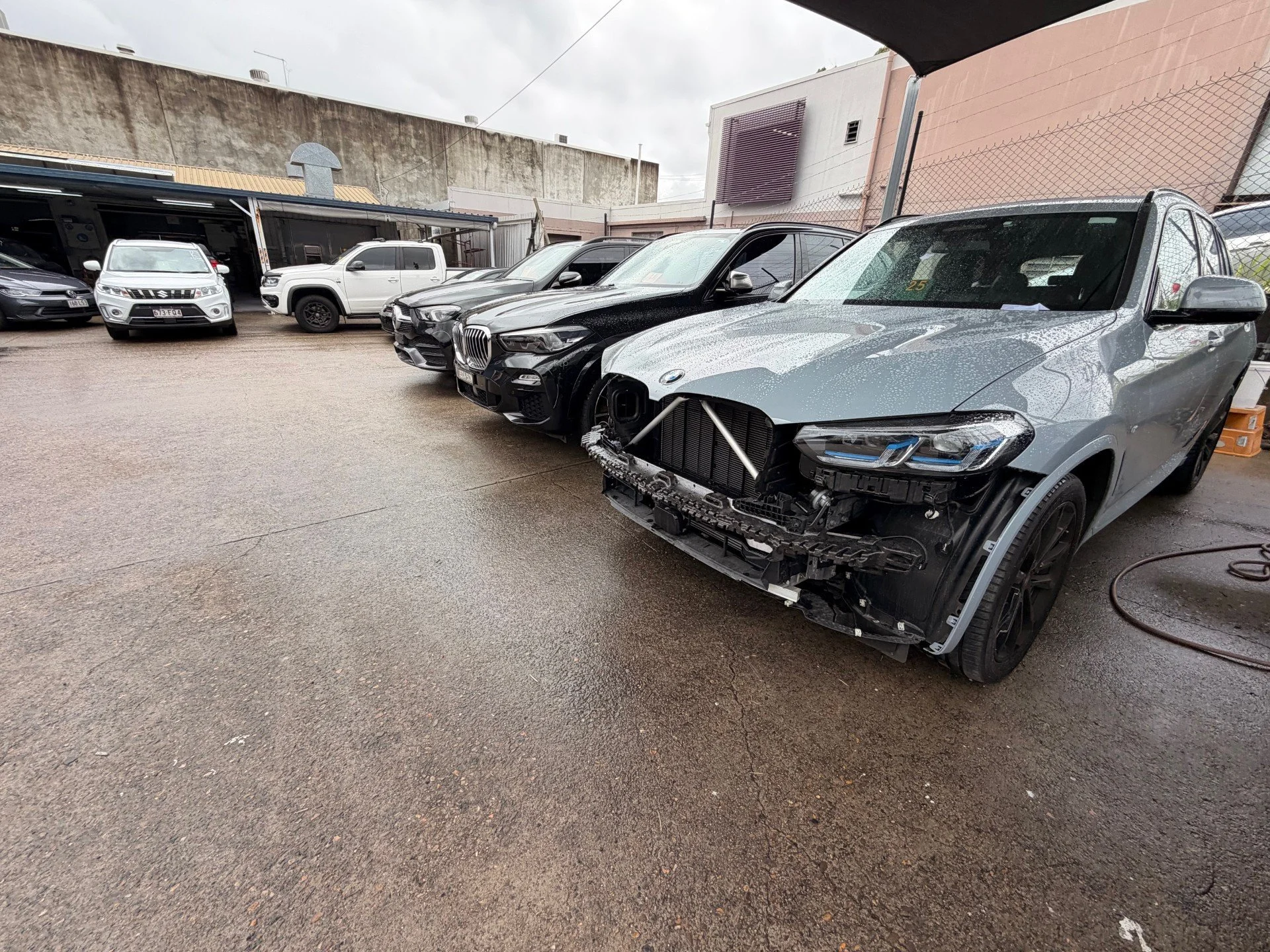 A row of cars parked outside at a car repair shop, with the front of a damaged silver BMW SUV in the foreground, missing its front bumper and grille.