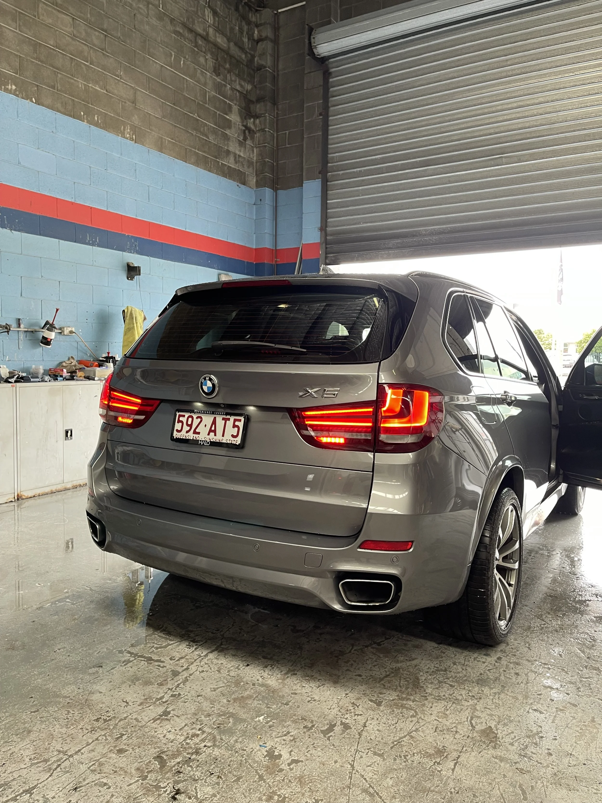 Gray BMW X6 inside an automotive workshop, partially inside a garage with a corrugated metal roll-up door, with tools on a workbench in the background.