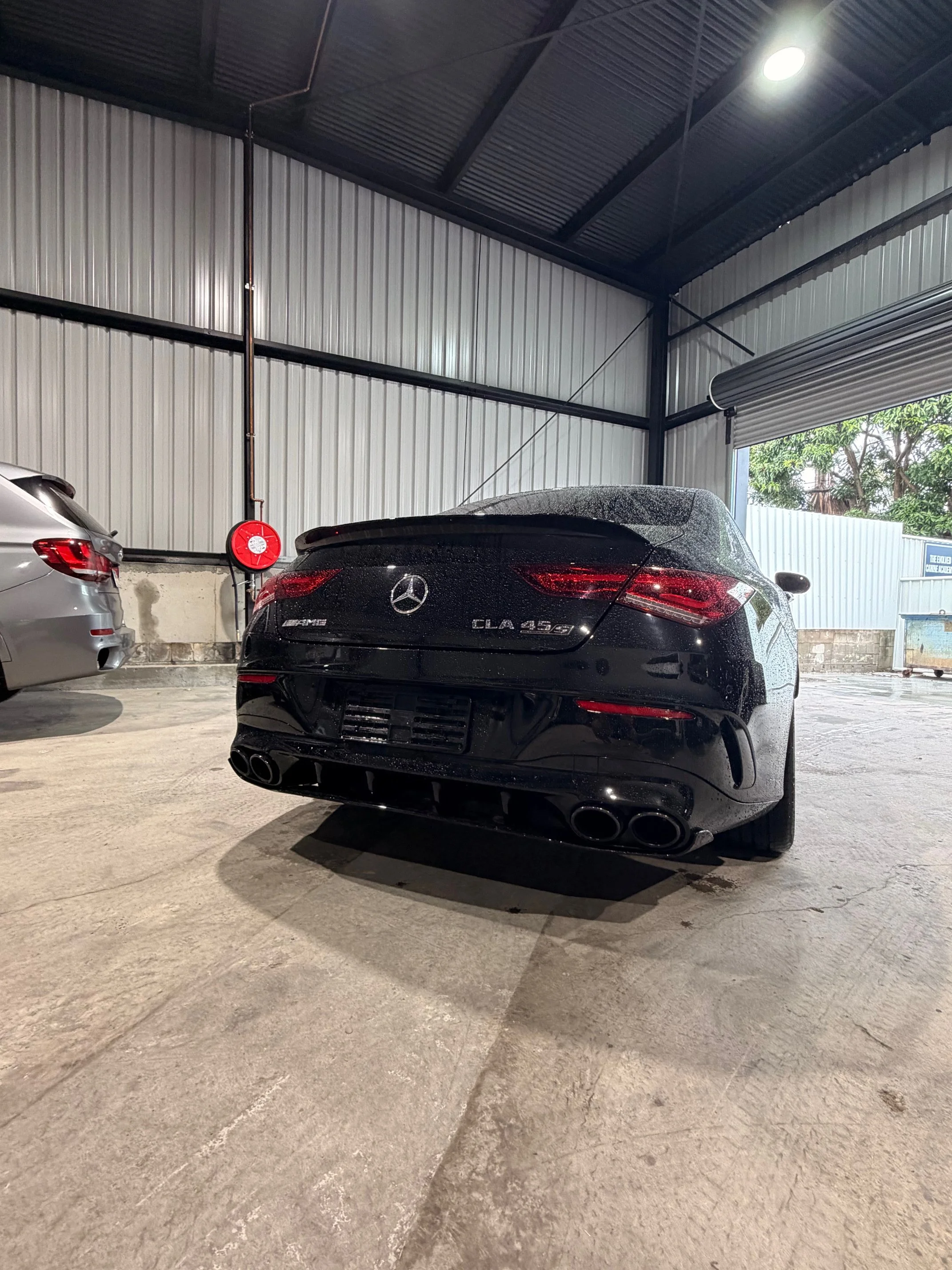 Black Mercedes-Benz CLA 45S parked inside a warehouse with a grey car on the left and a partially open roller door showing trees outside.
