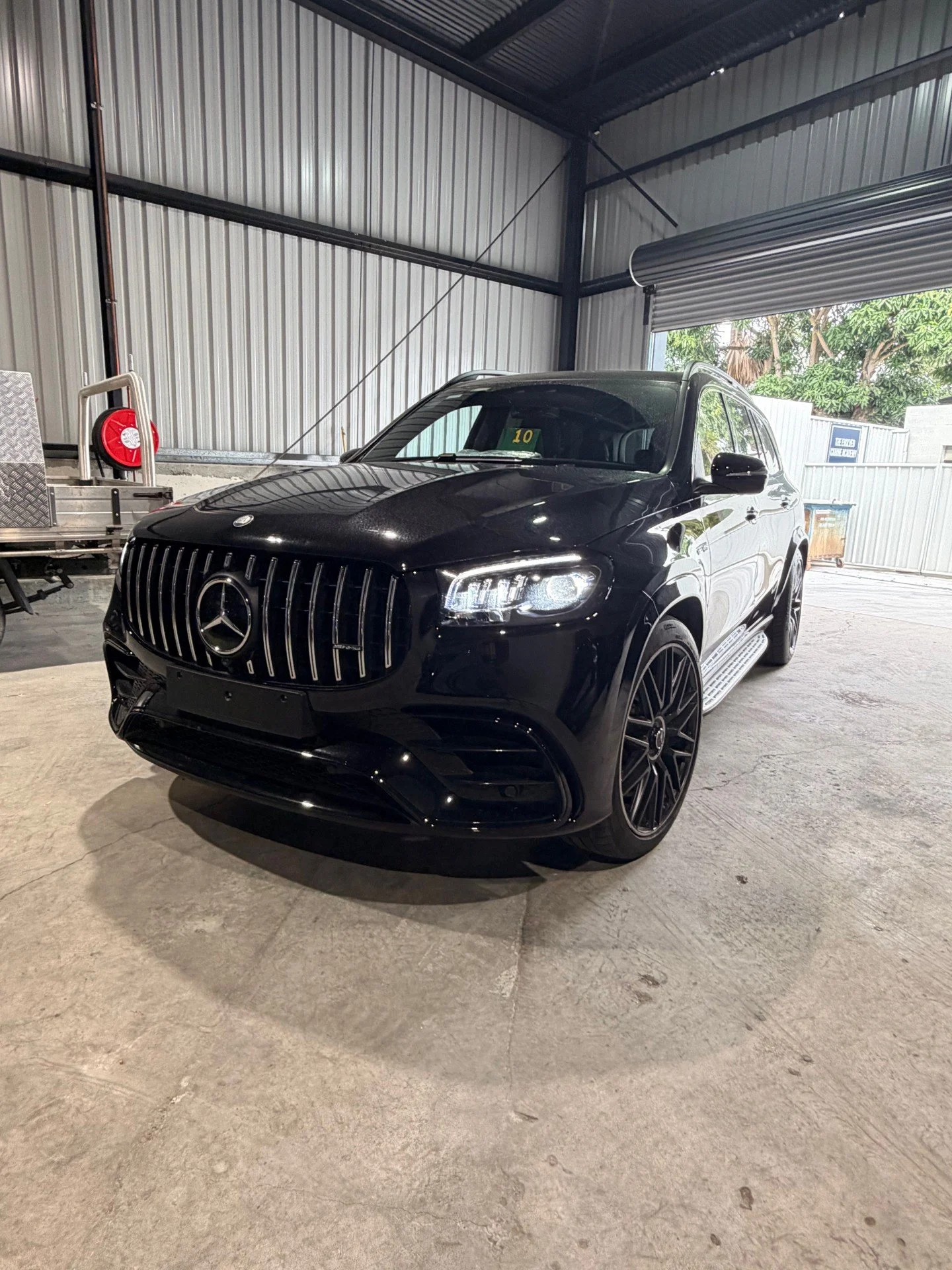 A black Mercedes-Benz SUV parked inside a garage with metal walls and ceiling, with a rolling garage door open to the outside where green trees are visible.