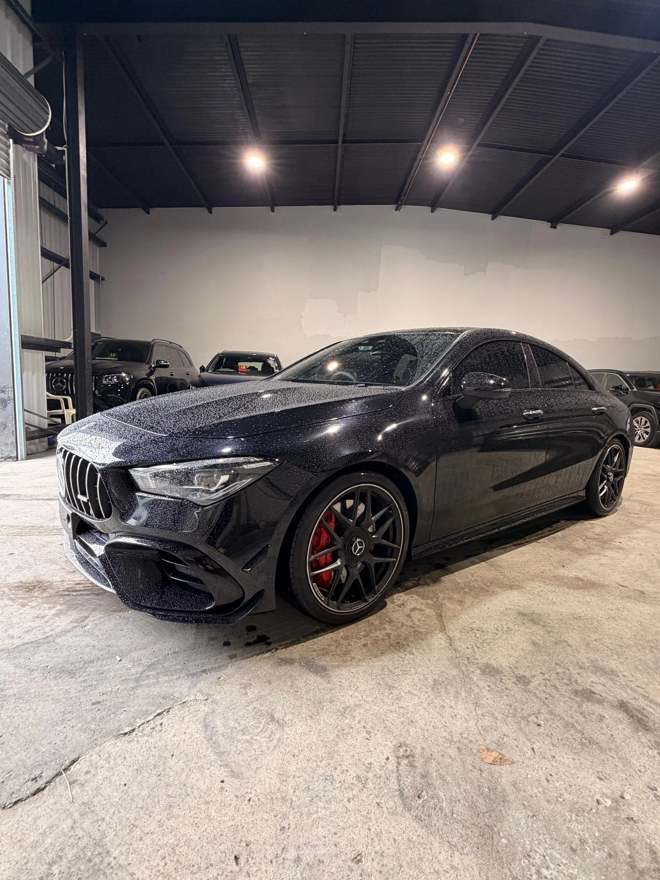 Black Mercedes-Benz sedan with tinted windows, red brake calipers, and a wet surface parked inside a warehouse or garage with other vehicles in the background.