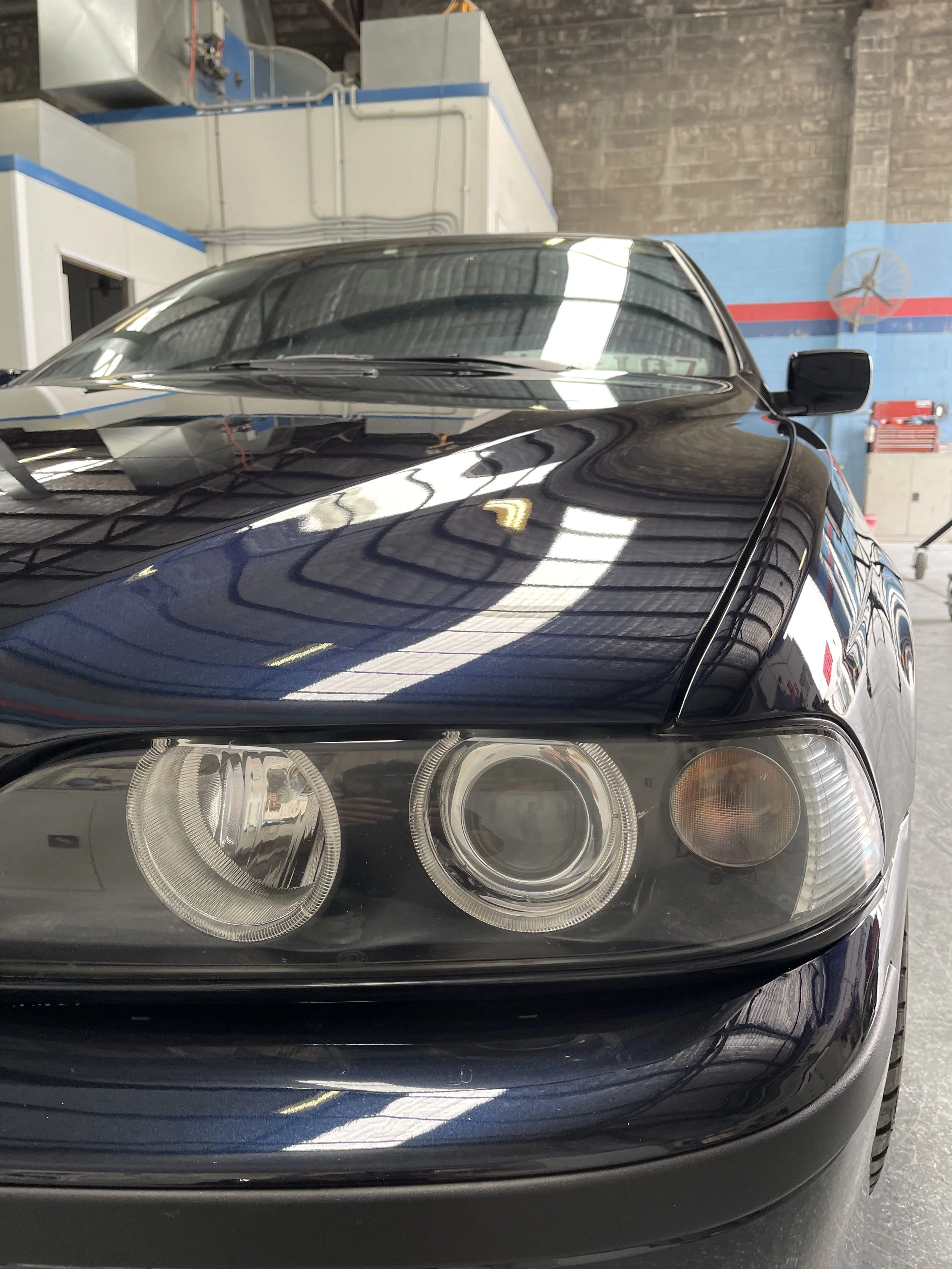 Close-up of the front quarter of a black sedan car parked inside a garage, showing the headlight, side mirror, and reflections on the hood and windshield.