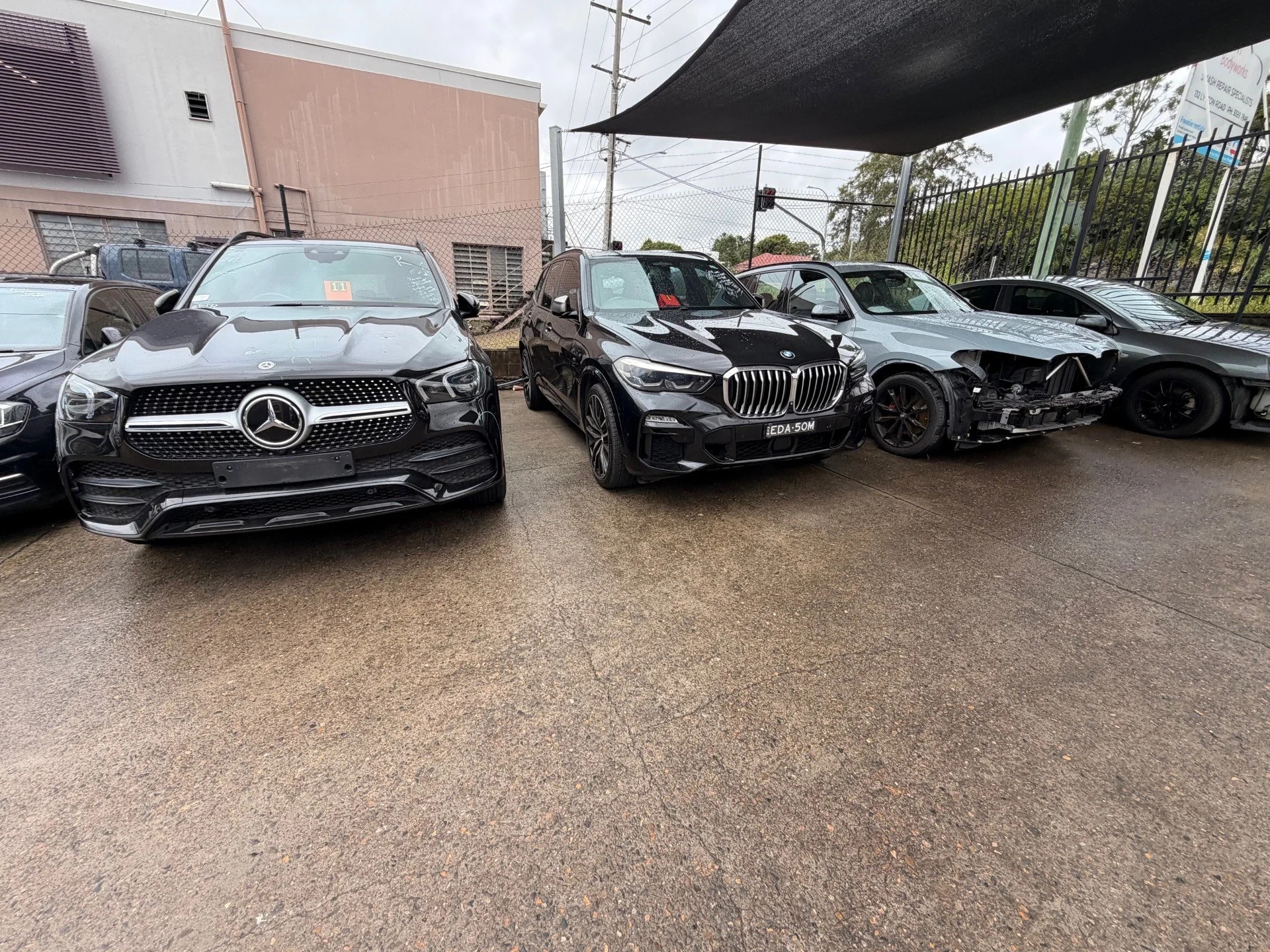 Four cars parked in a lot: a black Mercedes-Benz, a black BMW, a damaged gray car with front missing, and a gray car on the far right.