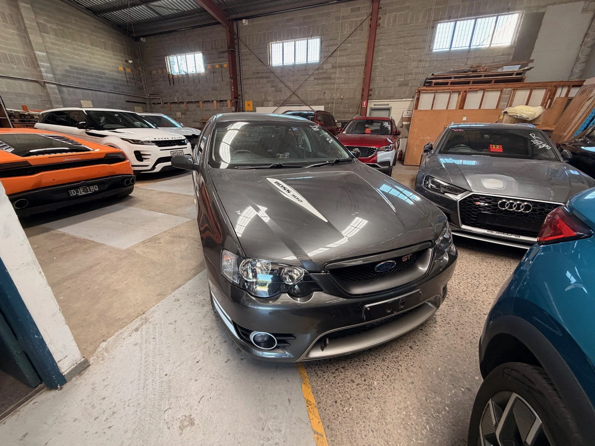 A collection of luxury cars parked inside a warehouse, featuring a gray Ford Focus ST in the foreground, a silver Audi, a white Range Rover, an orange Lamborghini, and a red car among others.