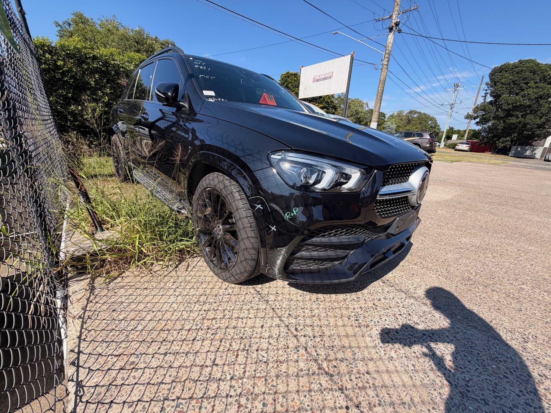Black Mercedes-Benz SUV parked next to a chain-link fence on a gravel lot under a clear blue sky with power lines overhead.