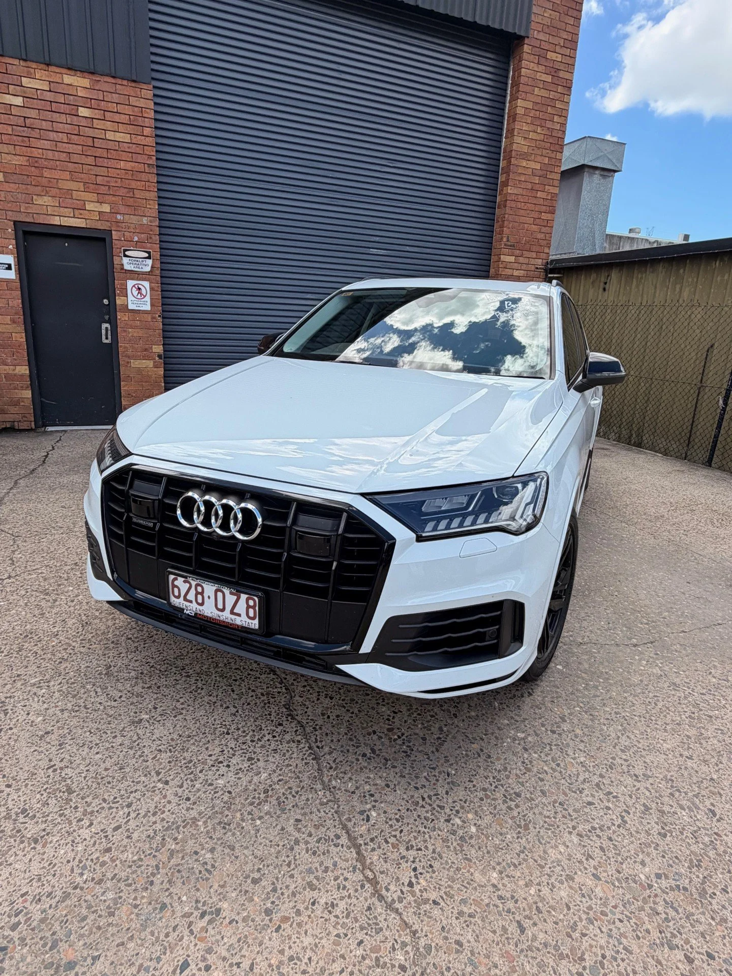 A white Audi SUV parked in front of a brick building with a black garage door and a small black door, reflecting the cloudy sky in its windshield.
