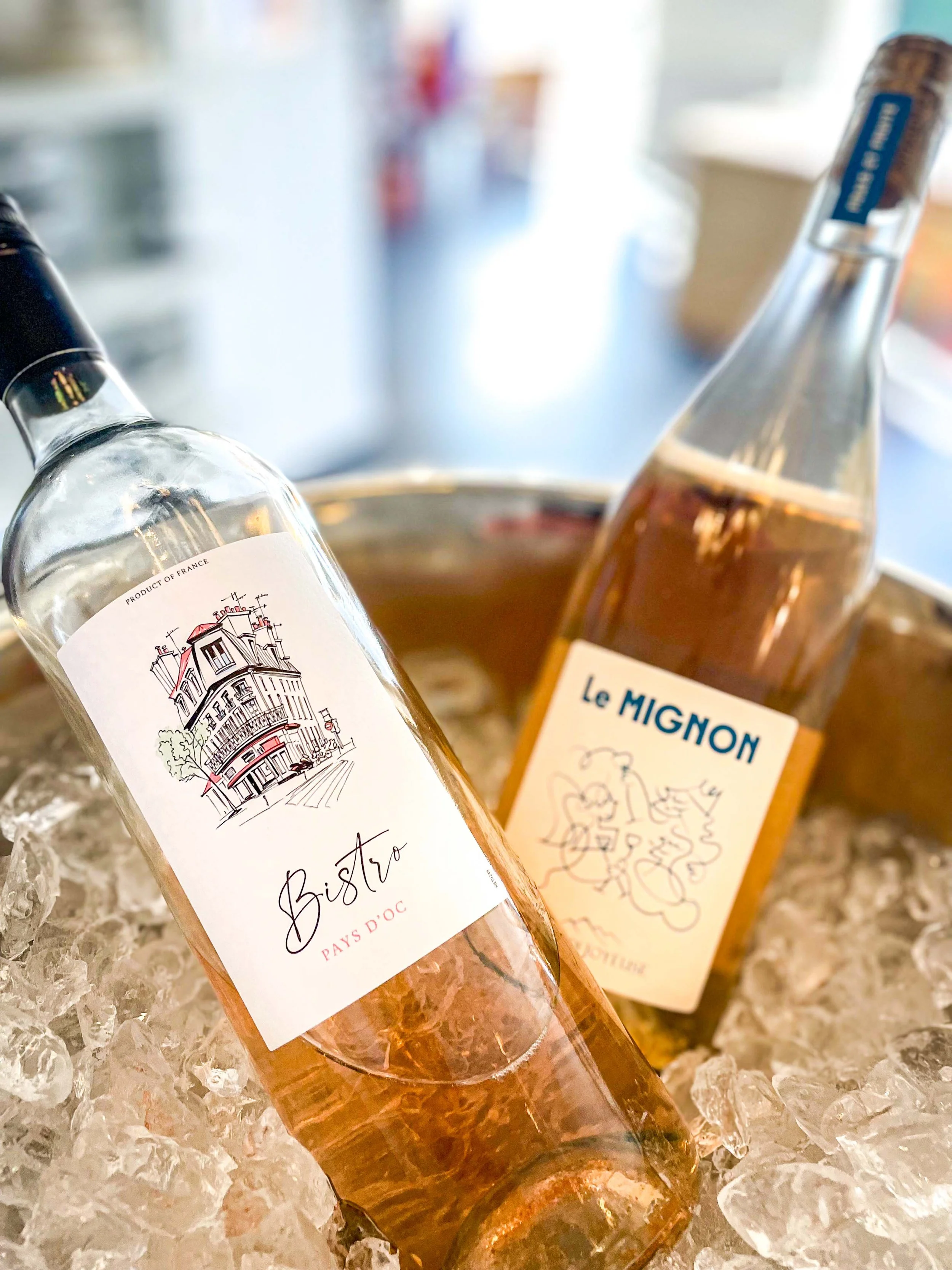 To flasker vin, en hvid og en rosé, ligger på is i en kurv. Flaskerne har labels med illustrationer af bygninger og vinøse beskrivelser.