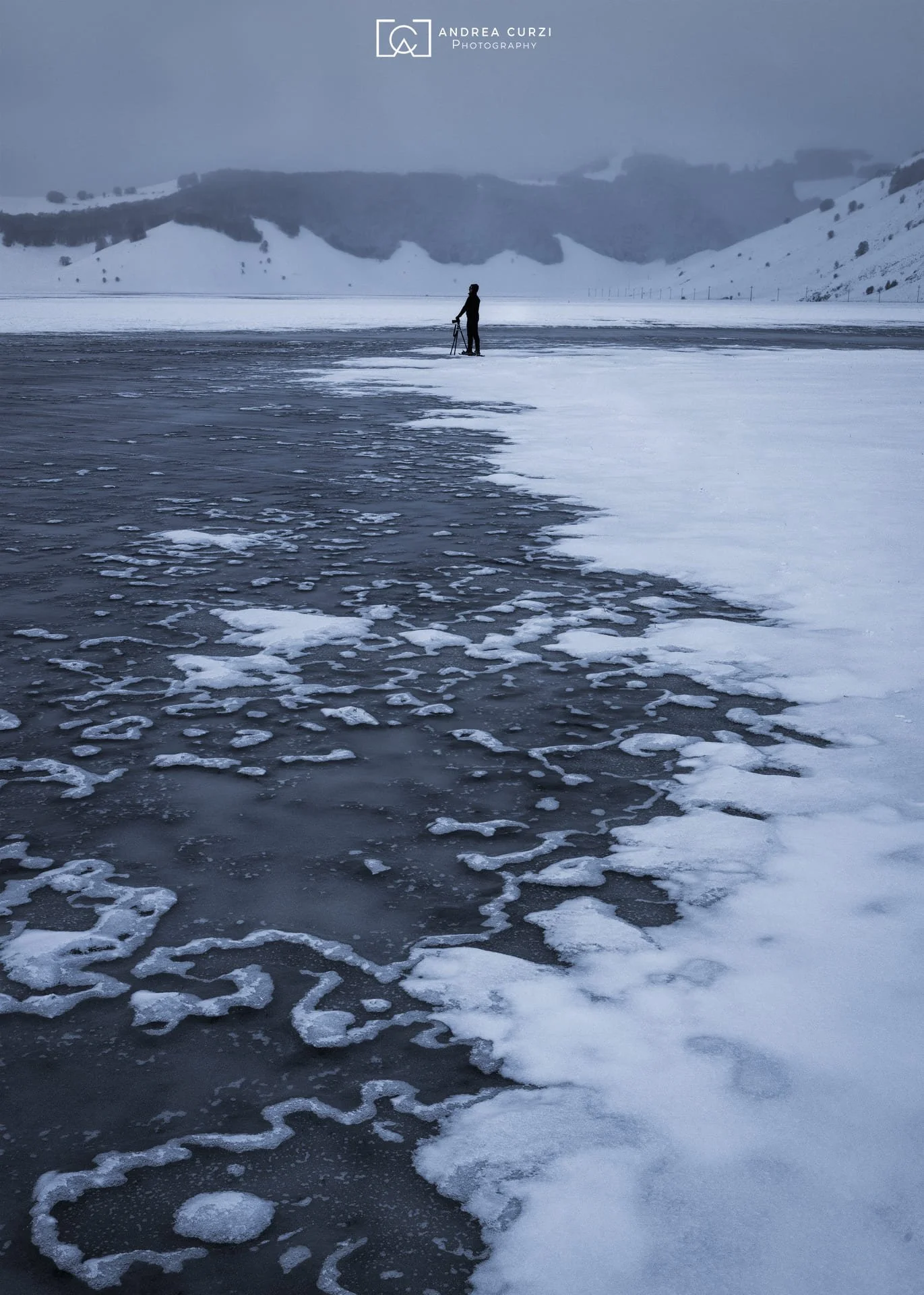 Una persona che cammina su un lago ghiacciato coperto di neve con uno sfondo di montagne e un cielo nuvoloso. Scattata sul Parco Nazionale dei Monti Sibillini a Castelluccio di Norcia durante un workshop fotografico di Andrea Curzi.