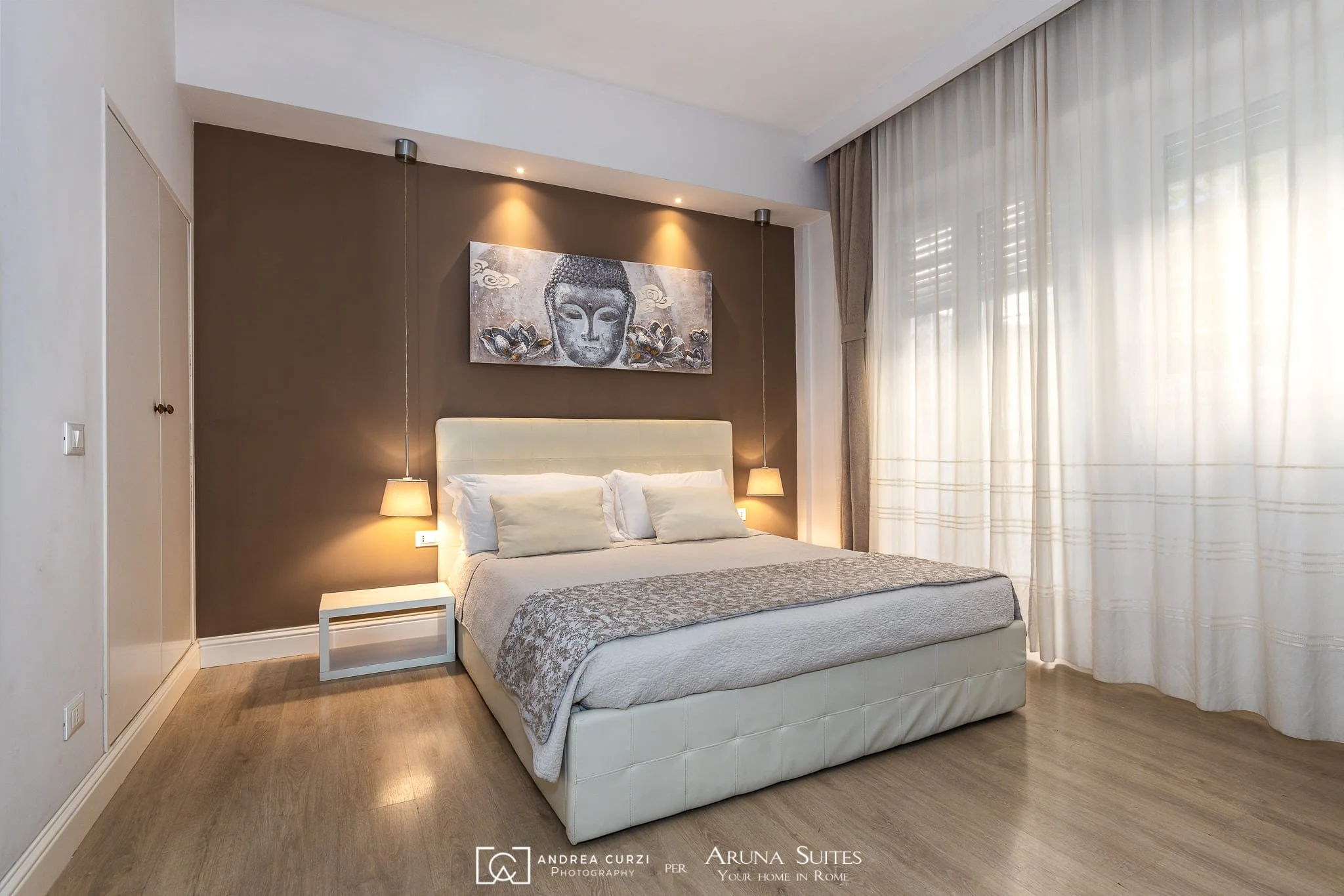 Camera da letto matrimoniale di un affittacamere, sopra dipinto di una testa di Buddha con fiori, divisa da tendaggi bianchi e grandi finestre. Scattata durante un servizio fotografico di interni di Andrea Curzi a Roma.